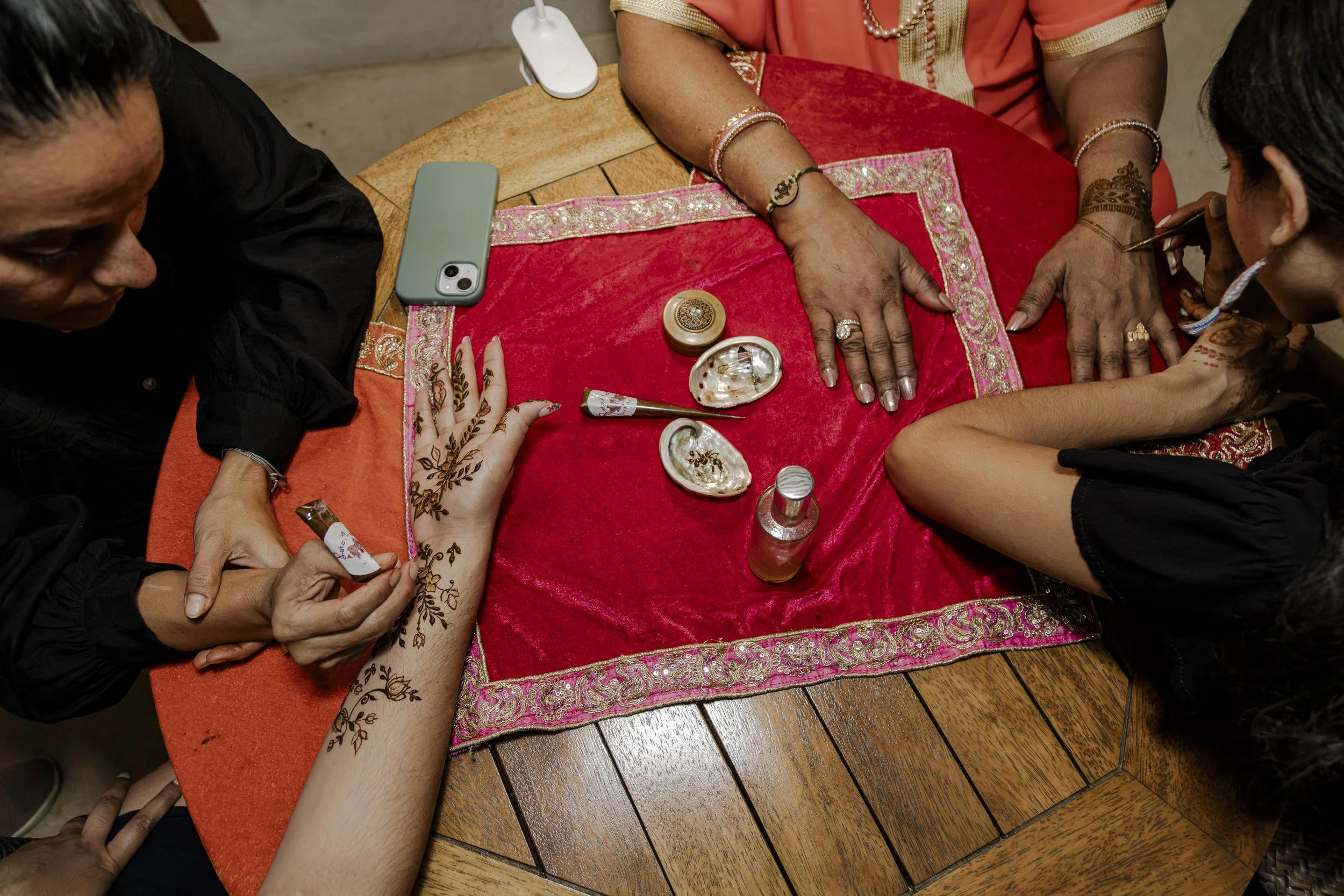 People getting henna designs applied on their hands at a table covered with a red cloth and various beauty supplies.