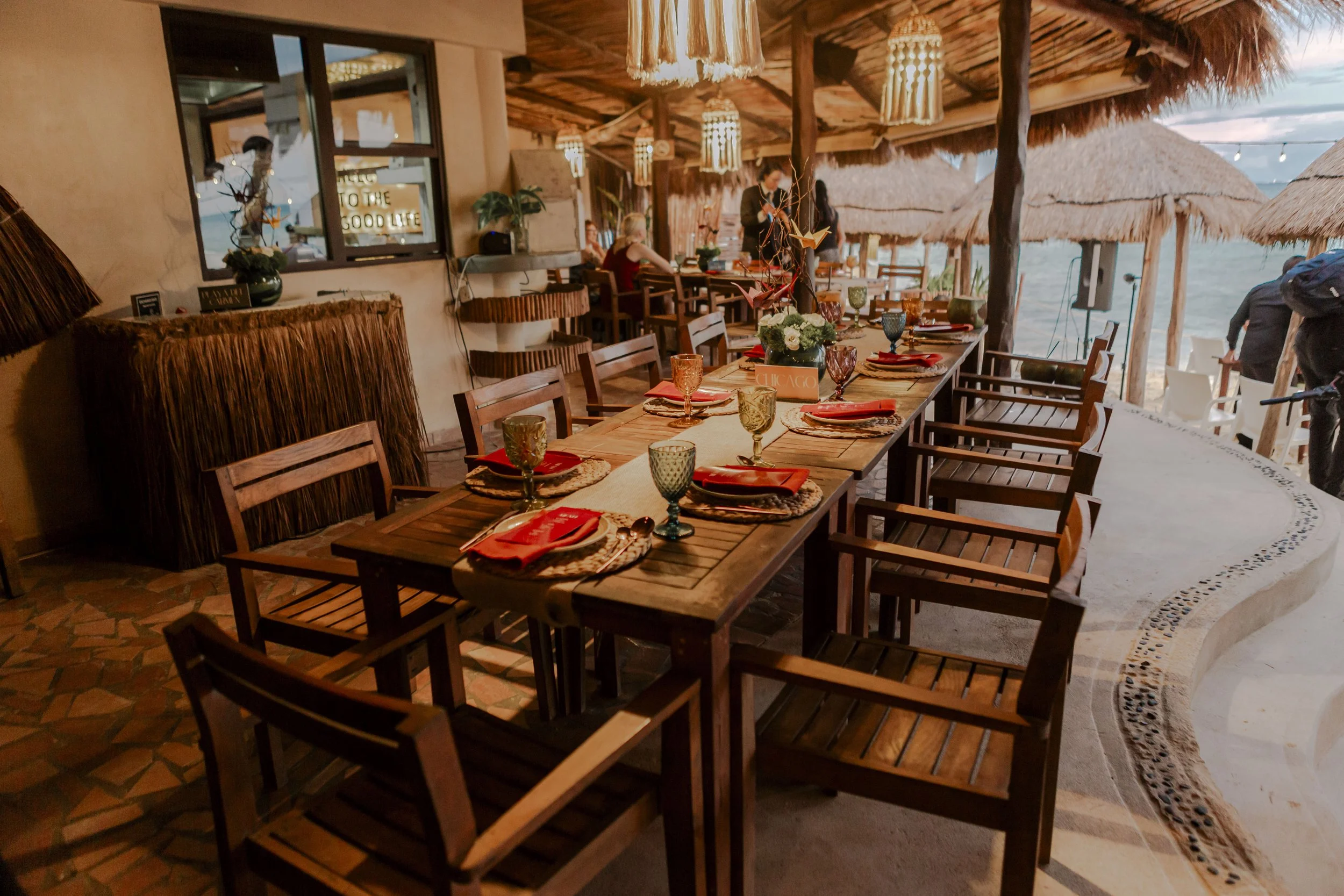 Beachside restaurant with a long wooden table set with colorful glassware and place settings, with a thatched roof and ocean in the background.