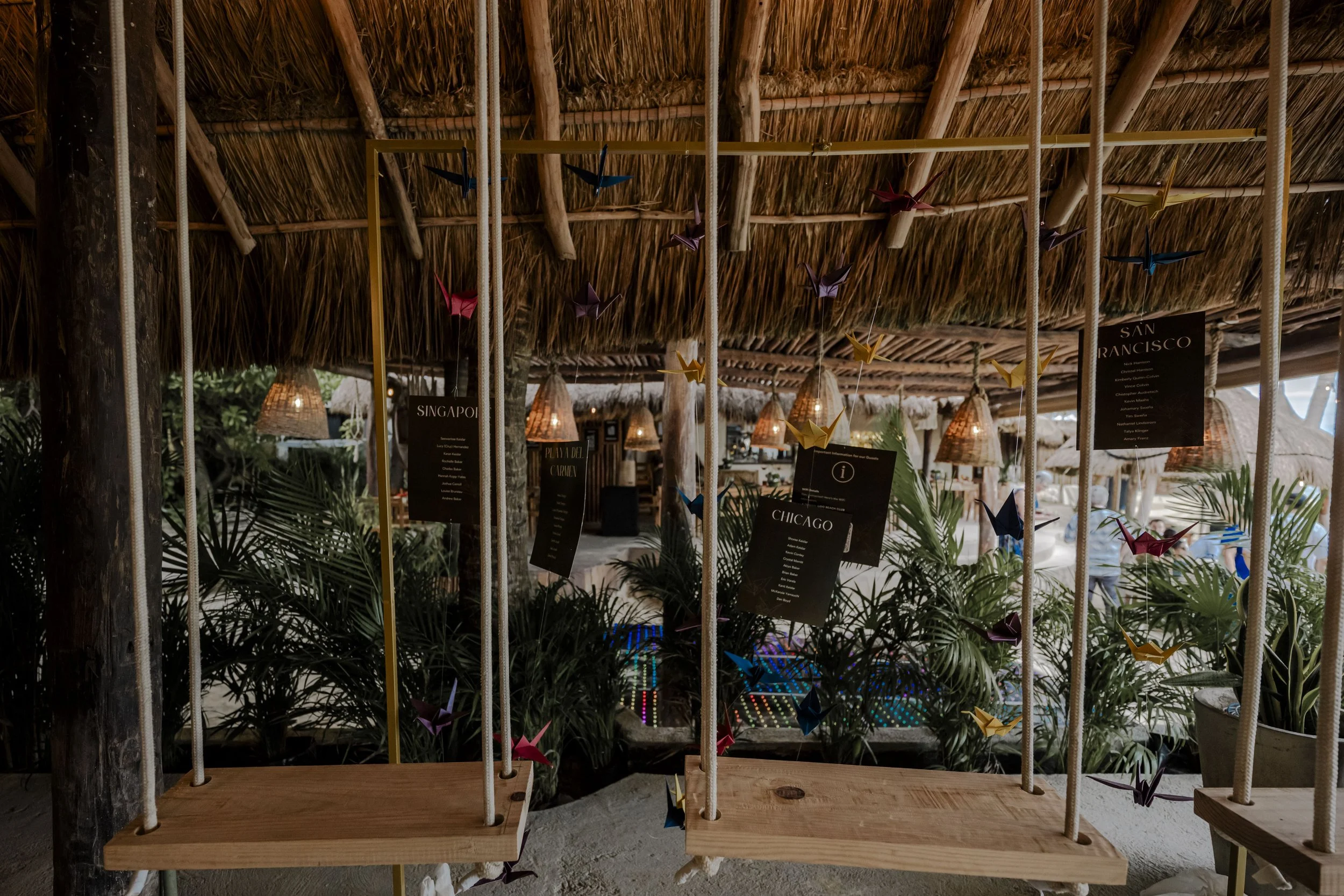 Decorative hanging swings with black event menus and colorful origami cranes inside a tiki hut with thatched roof and tropical plants.