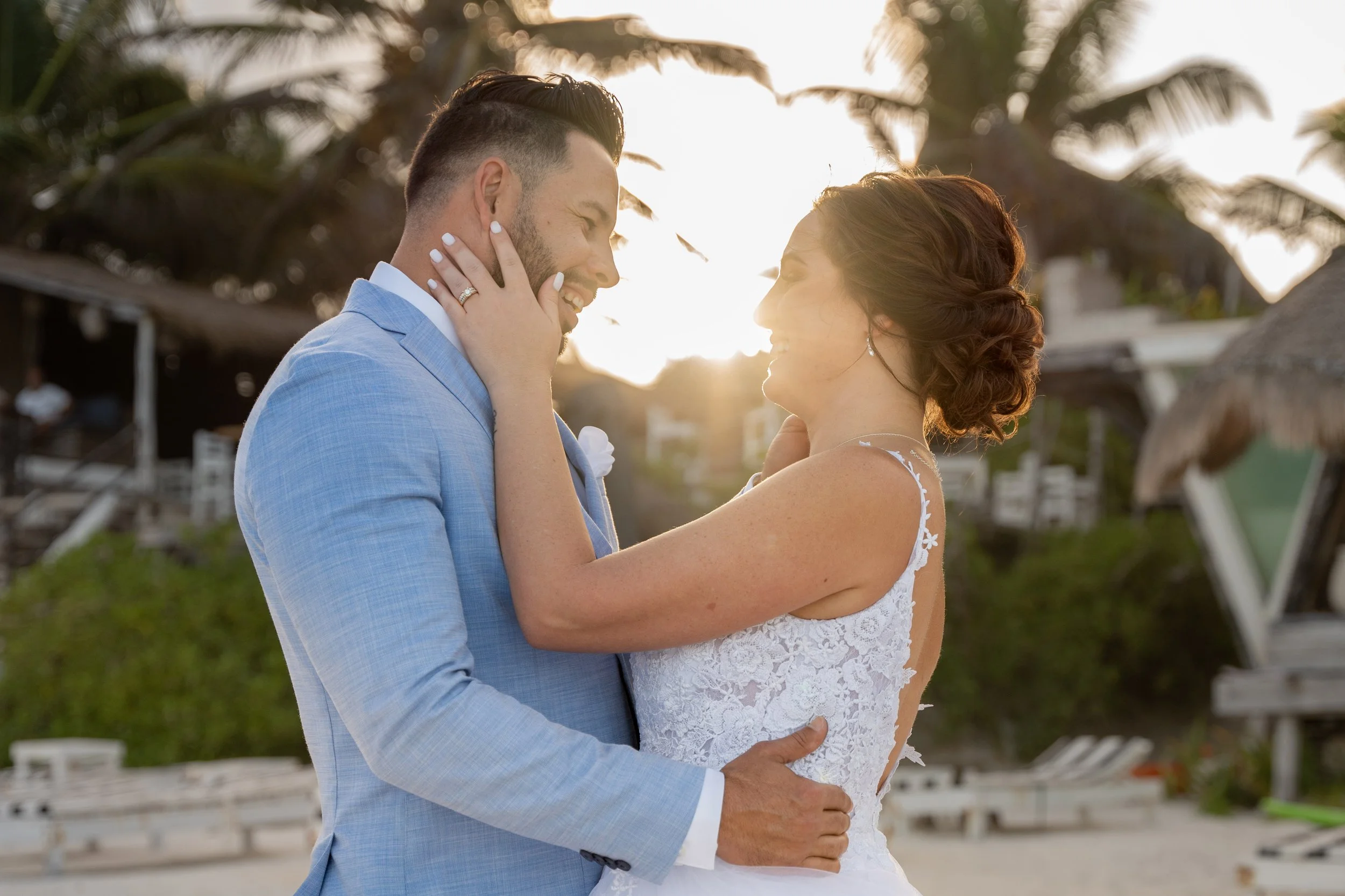 A newly married couple shares a romantic moment on the beach at sunset, with palm trees and beach chairs in the background.