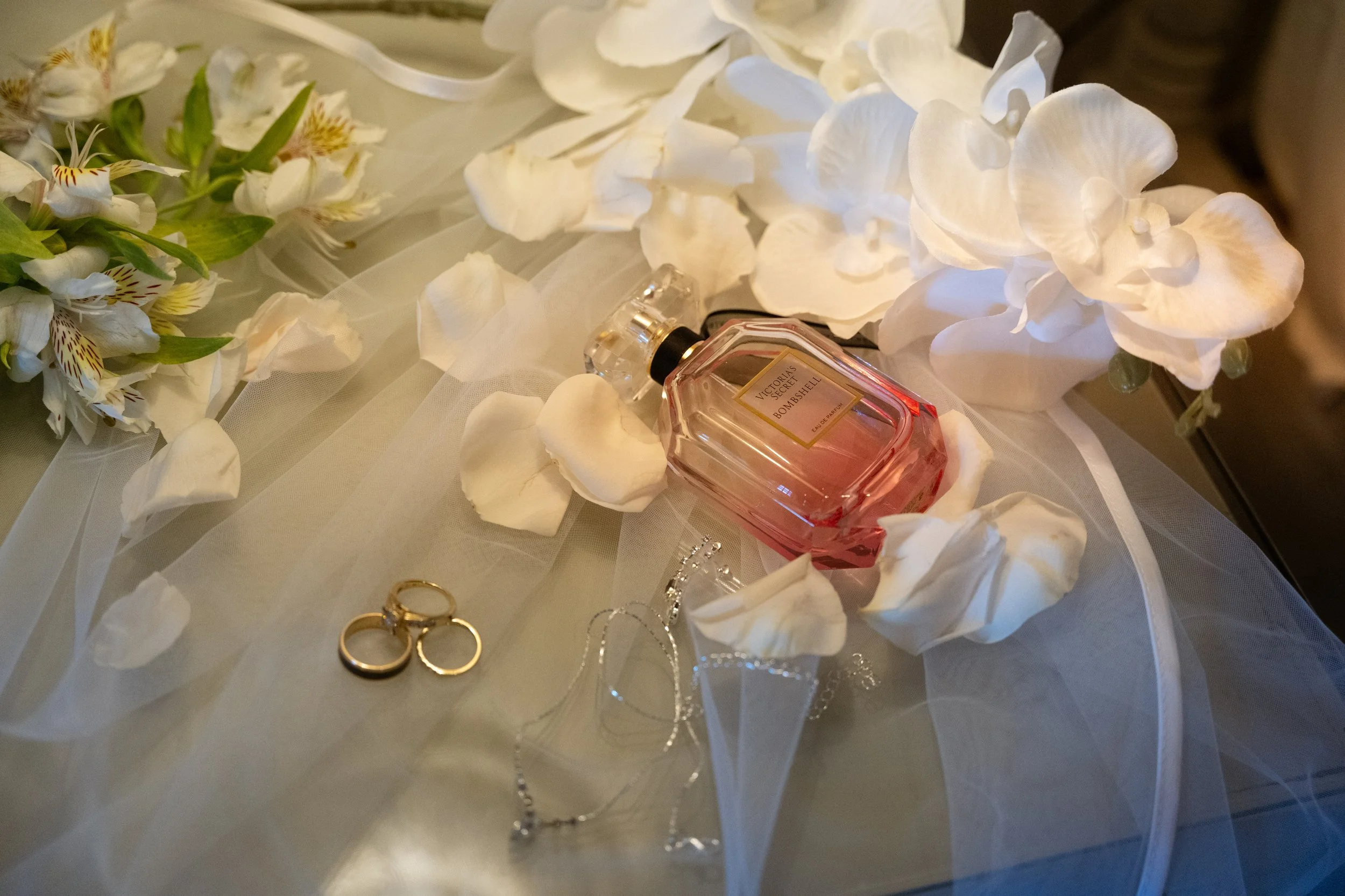 A bottle of Victoria's Secret Bombshell perfume, two gold wedding rings, and silver jewelry arranged on a surface with white flower petals and tulle fabric, possibly at a wedding or romantic event.