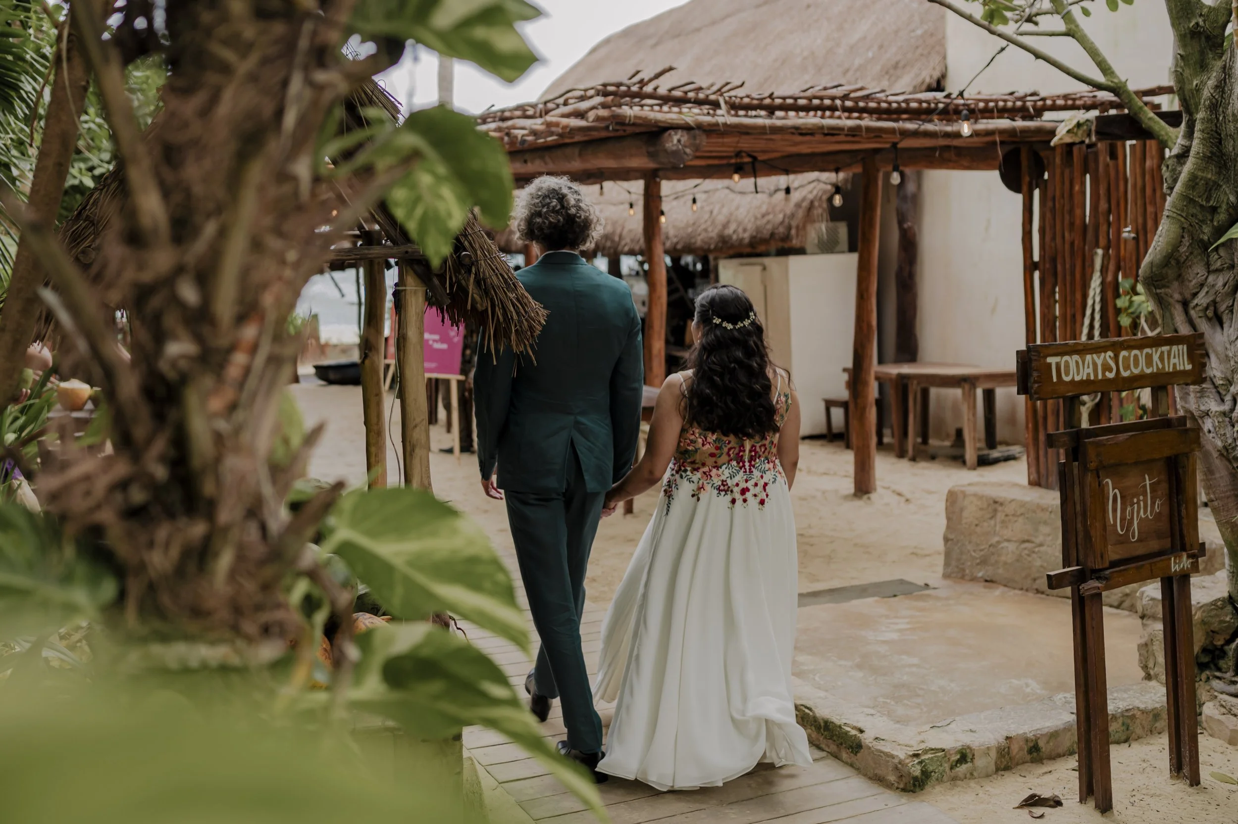 A couple holding hands walking on a sandy path towards a rustic outdoor bar area, with a wooden sign that reads 'Today's Cocktail'.