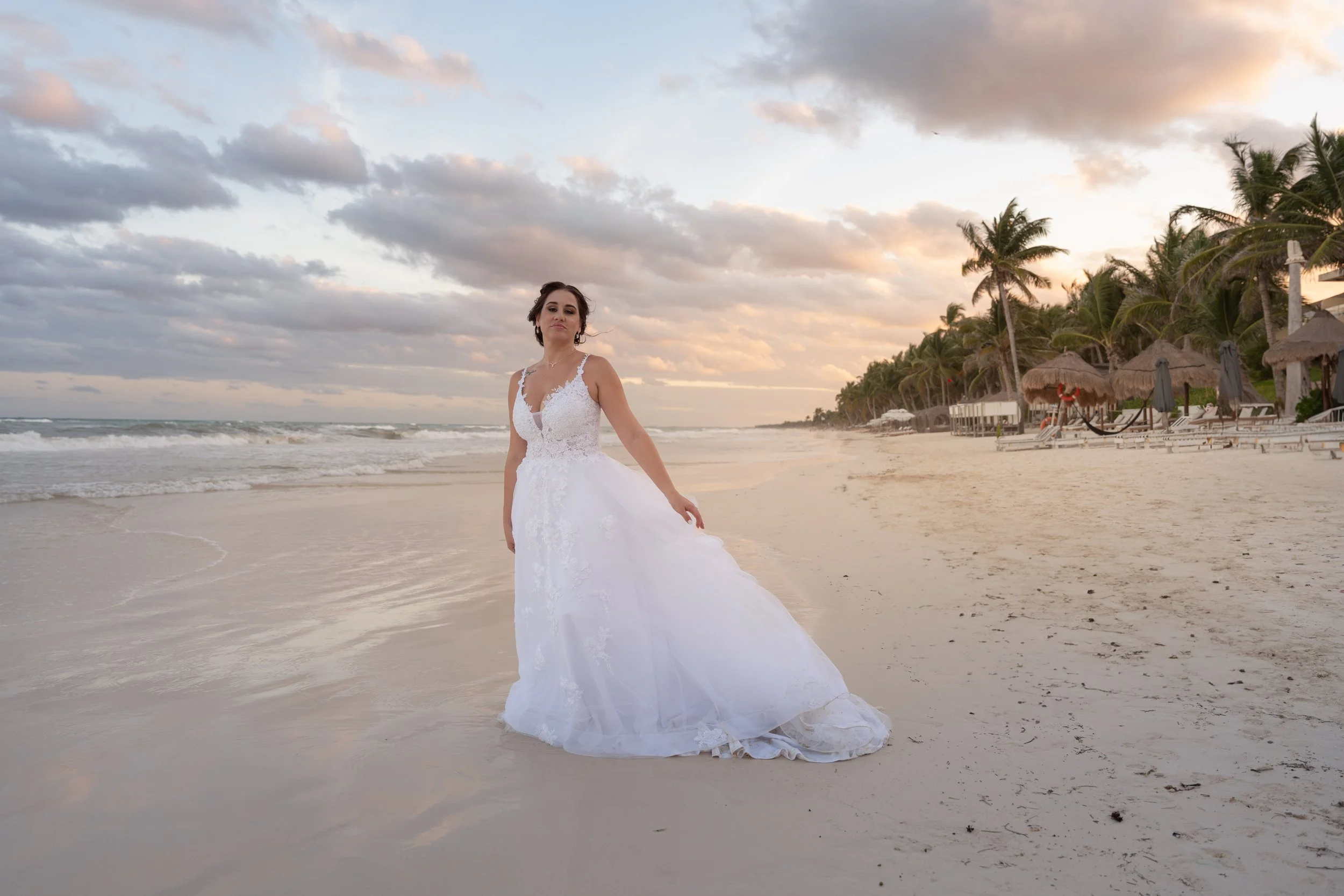 A woman in a white wedding dress standing on a sandy beach at sunset with palm trees and beach huts in the background.