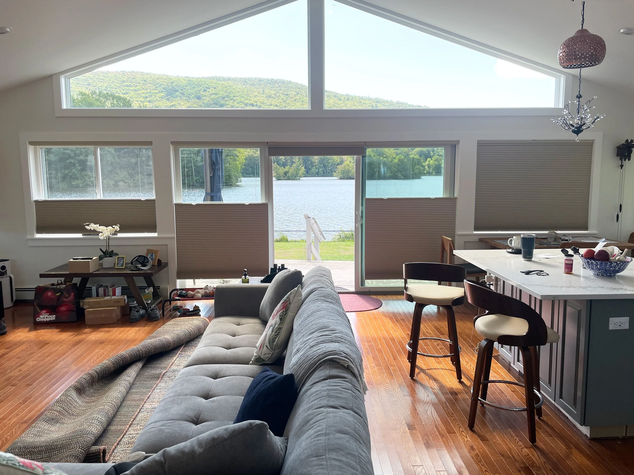 Living room with large windows showing a lake and hills outside, a sofa with pillows, a kitchen island with stools, and various items on the counter and floor.