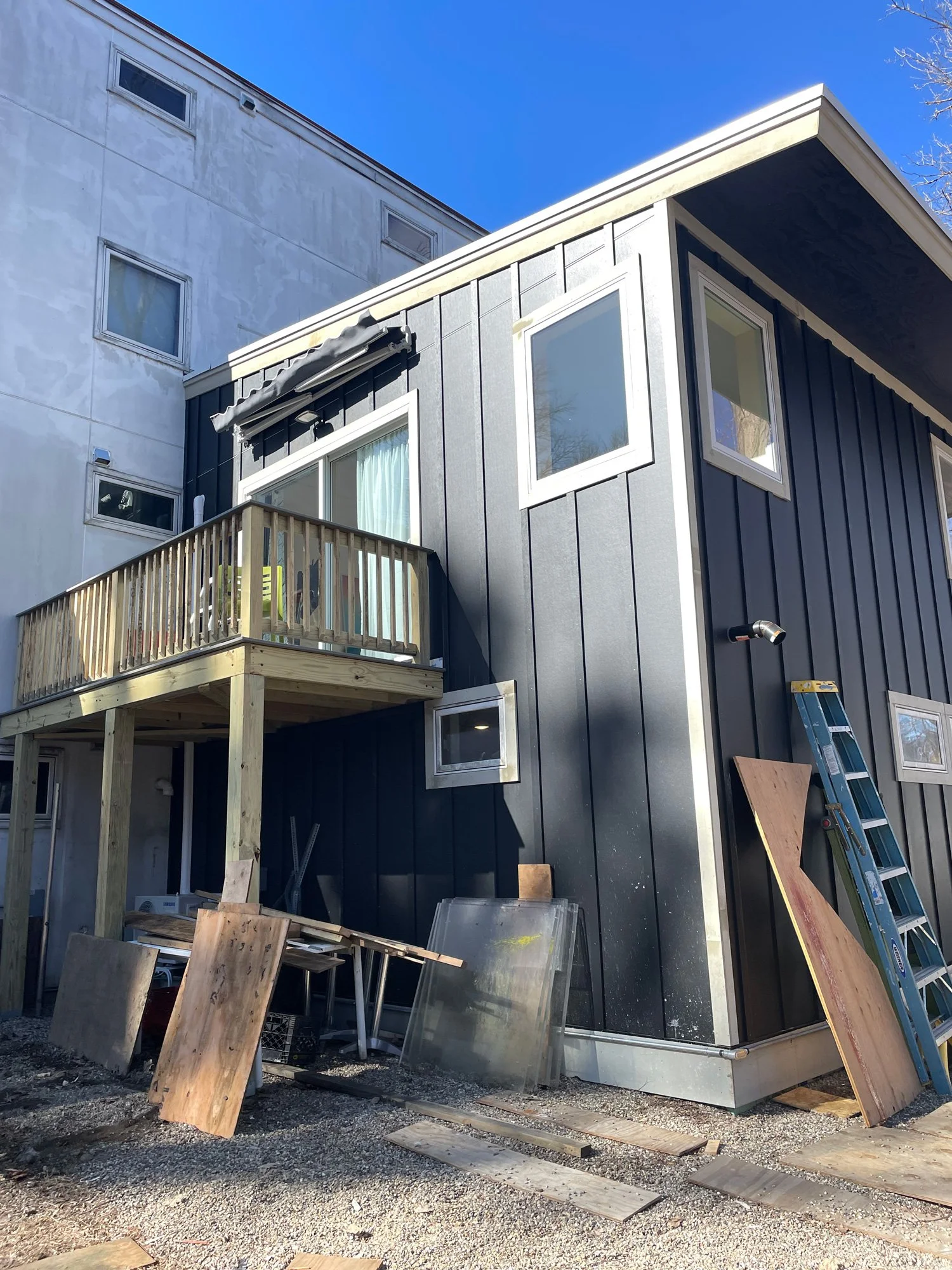 Construction site of a modern black house with a small balcony, a ladder, and building materials on the ground, under a clear blue sky.