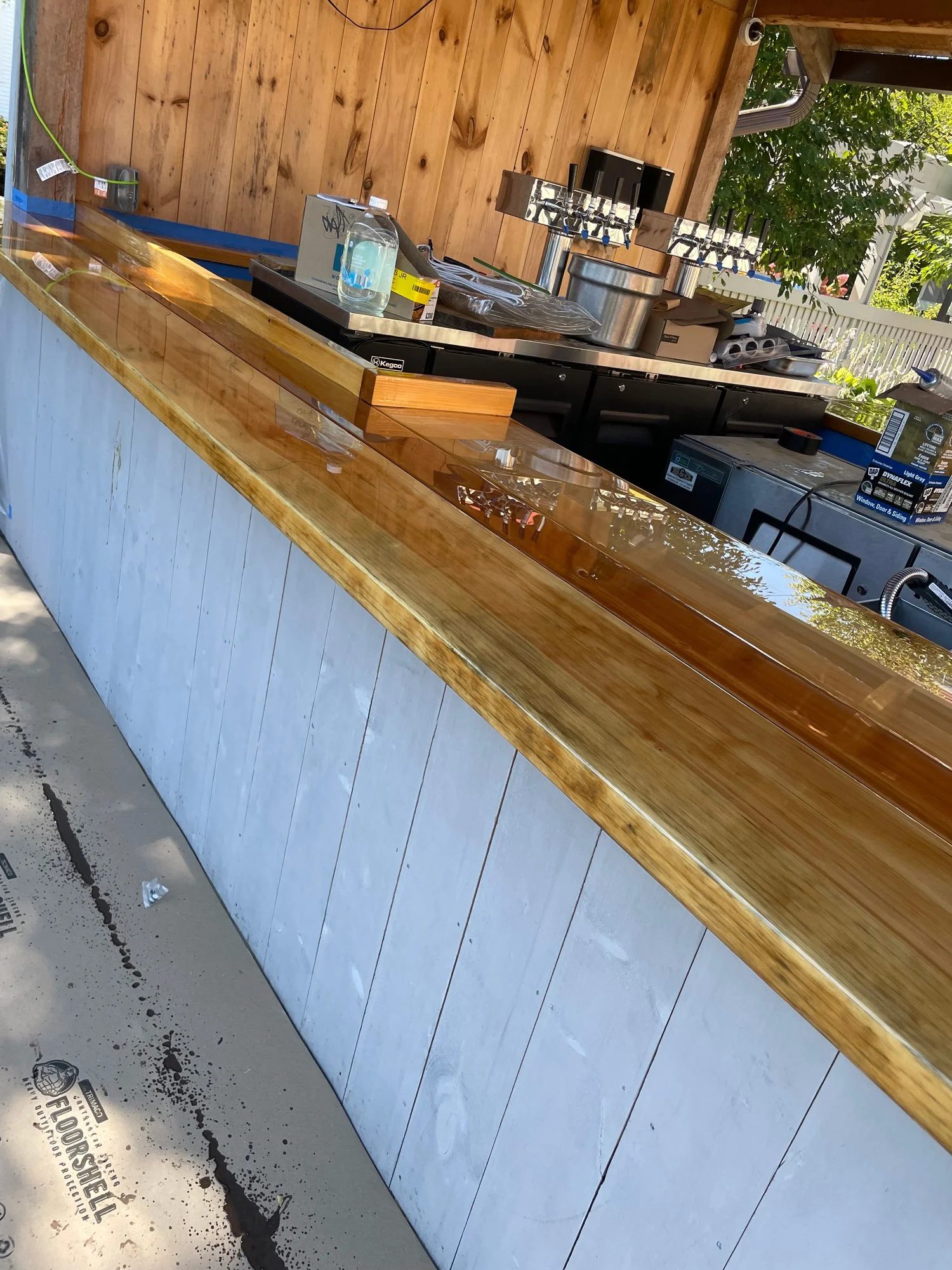 Construction of an outdoor bar area with a wooden countertop, tools, and equipment, with a wooden paneled wall in the background and a tree visible outside.