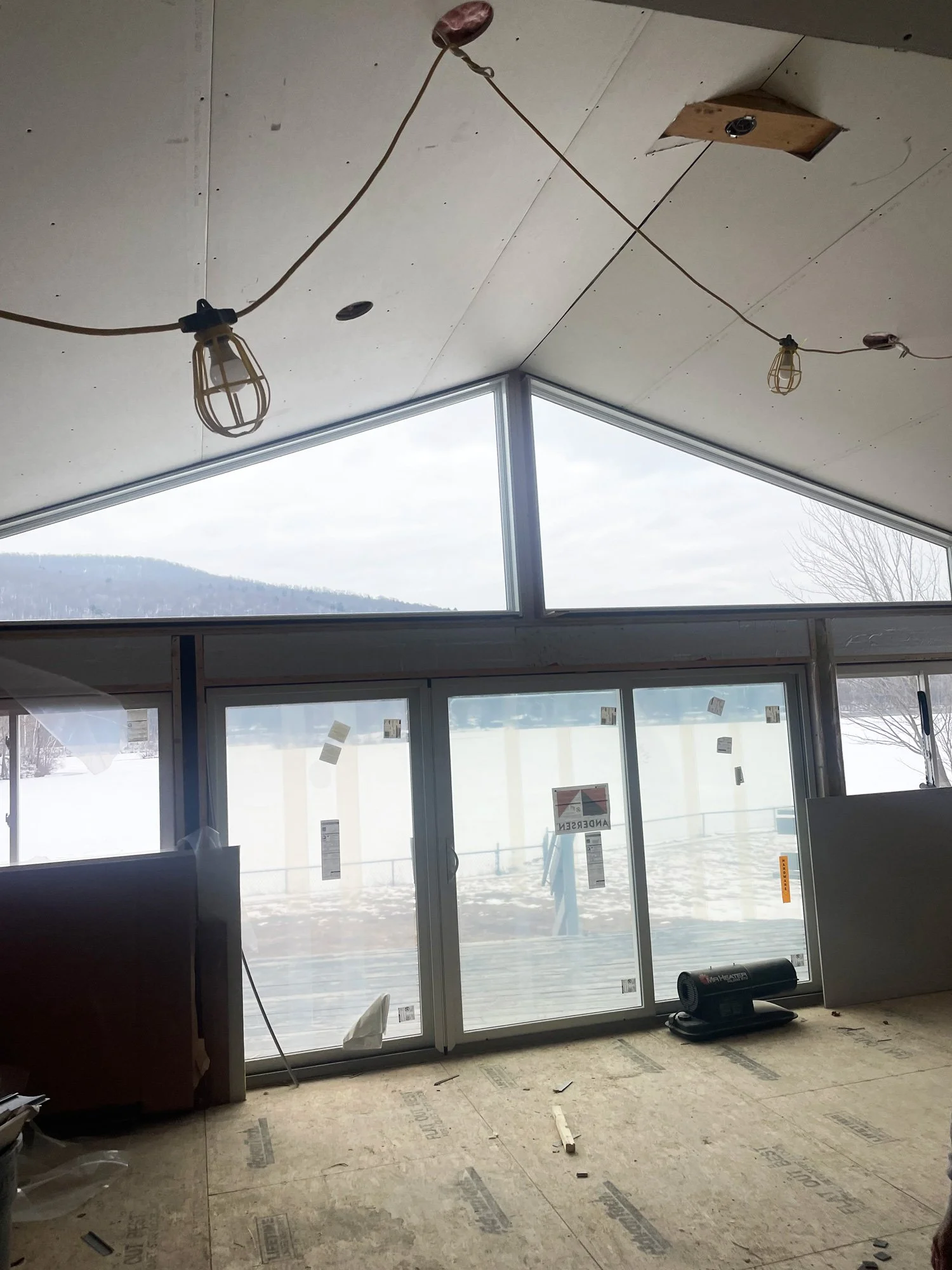Interior of a room under construction with large windows and a vaulted ceiling, light fixtures hanging from electrical cords, and construction materials on the floor.