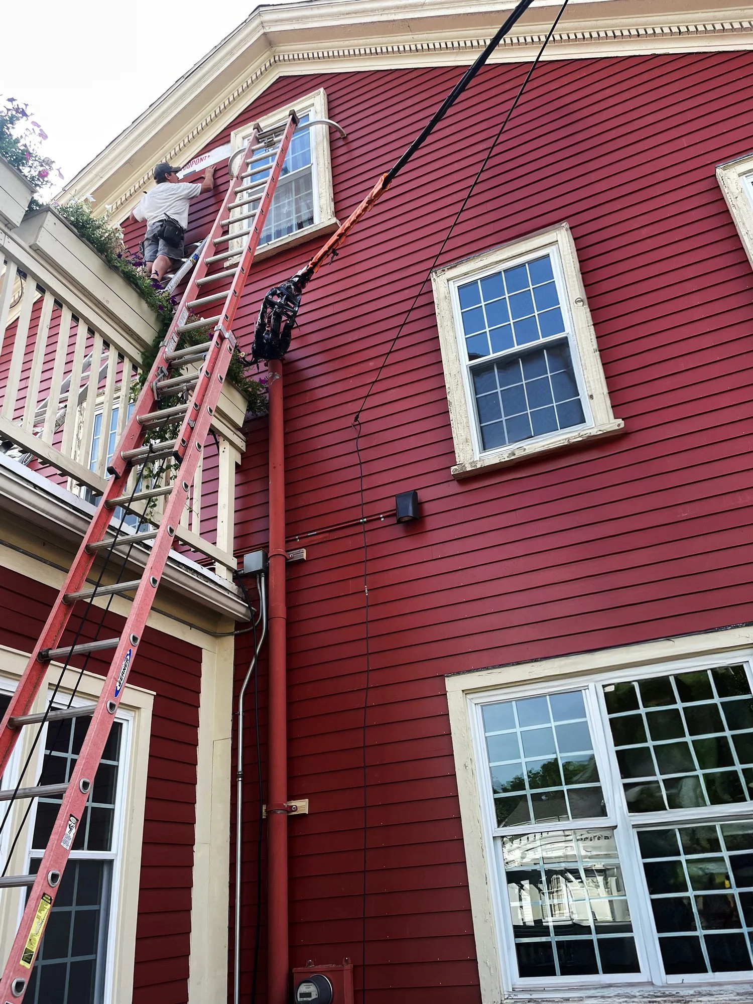 A person standing on a ladder on a red house, painting or repairing a window with white trim.