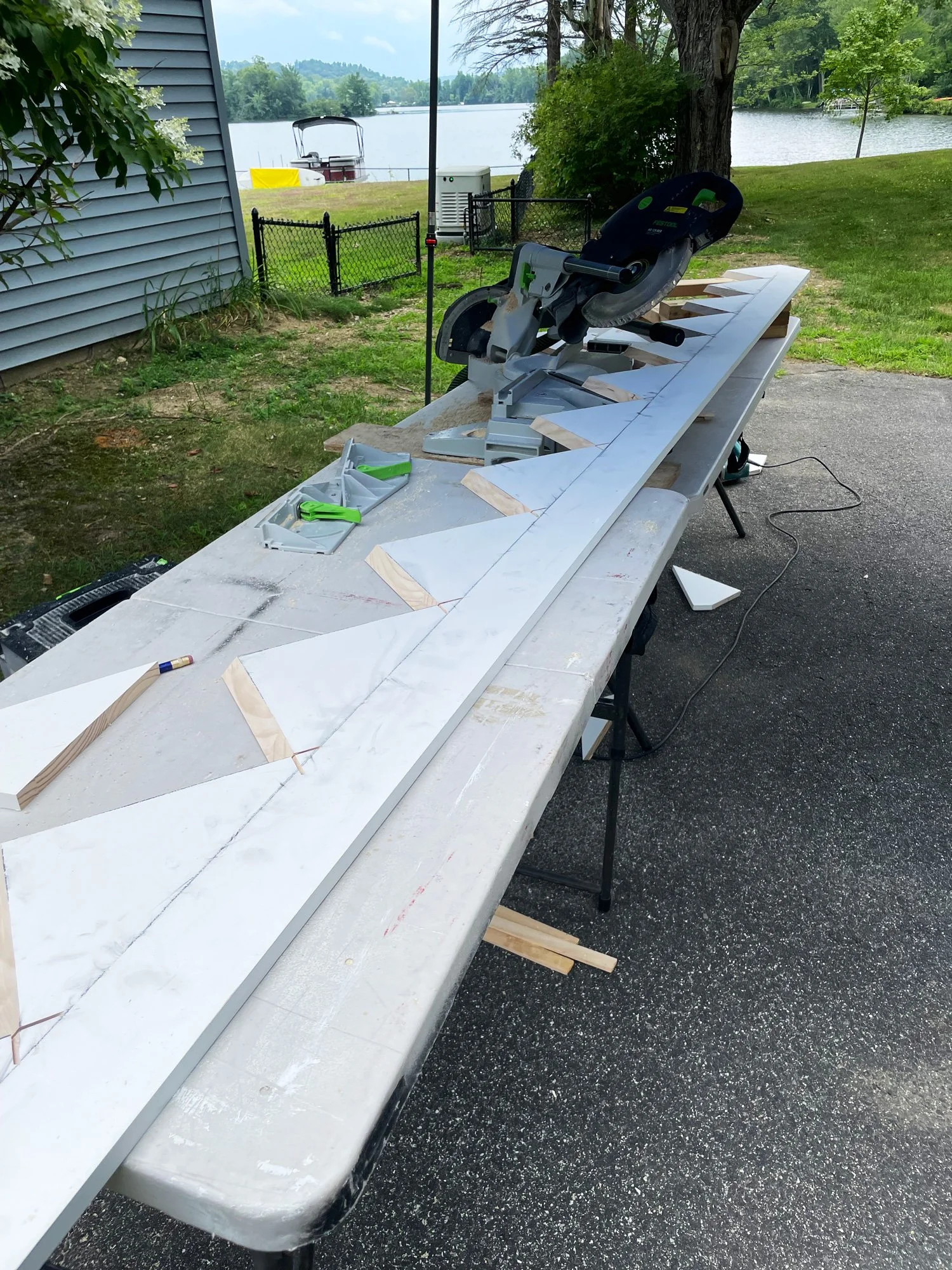 Outdoor workspace with a saw and wooden pieces on a table, near a lake, with trees and grass in the background.