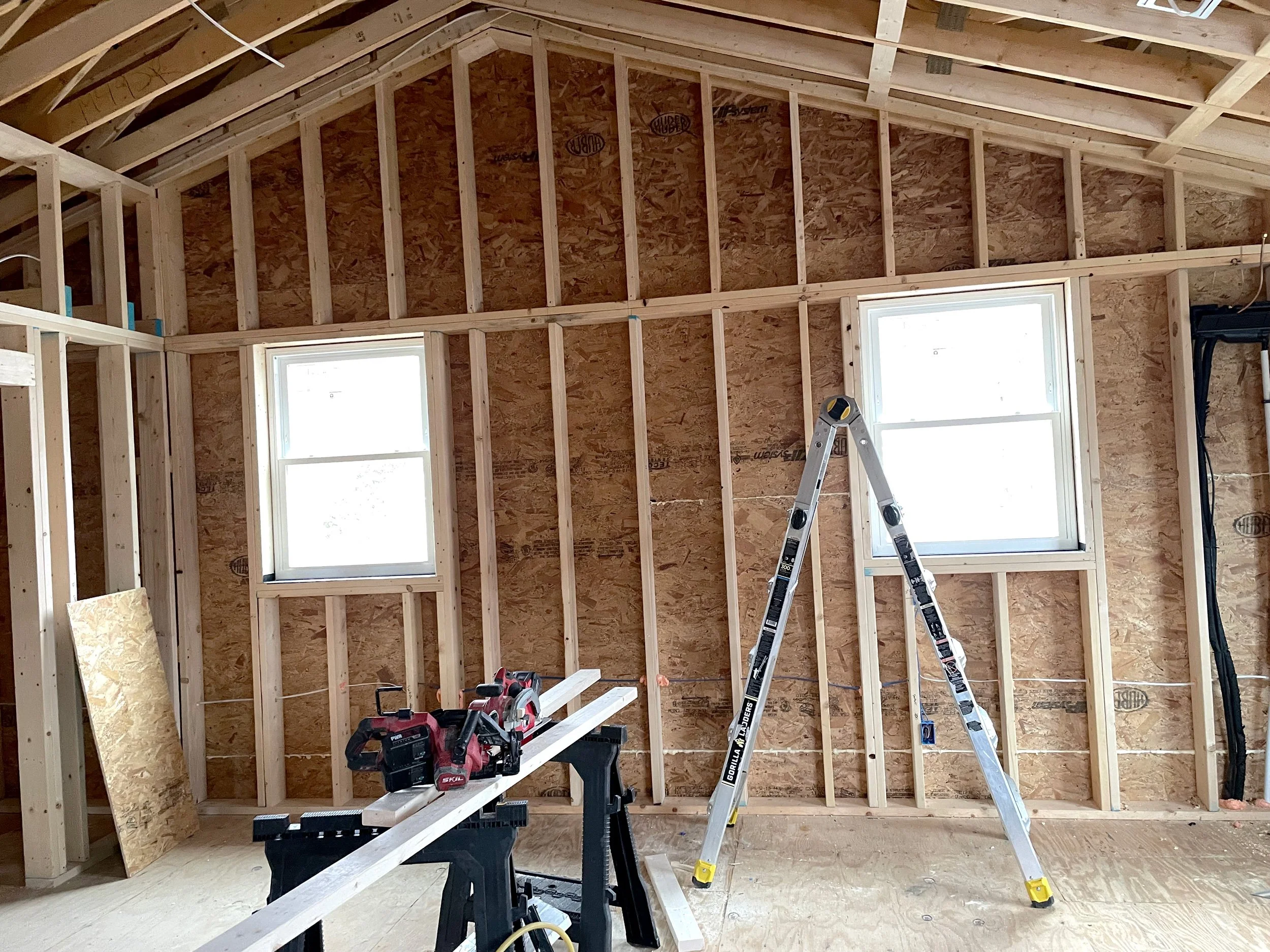 Interior of a house under construction with exposed wooden framing, two windows, and construction tools like a saw, ladder, and wooden planks.