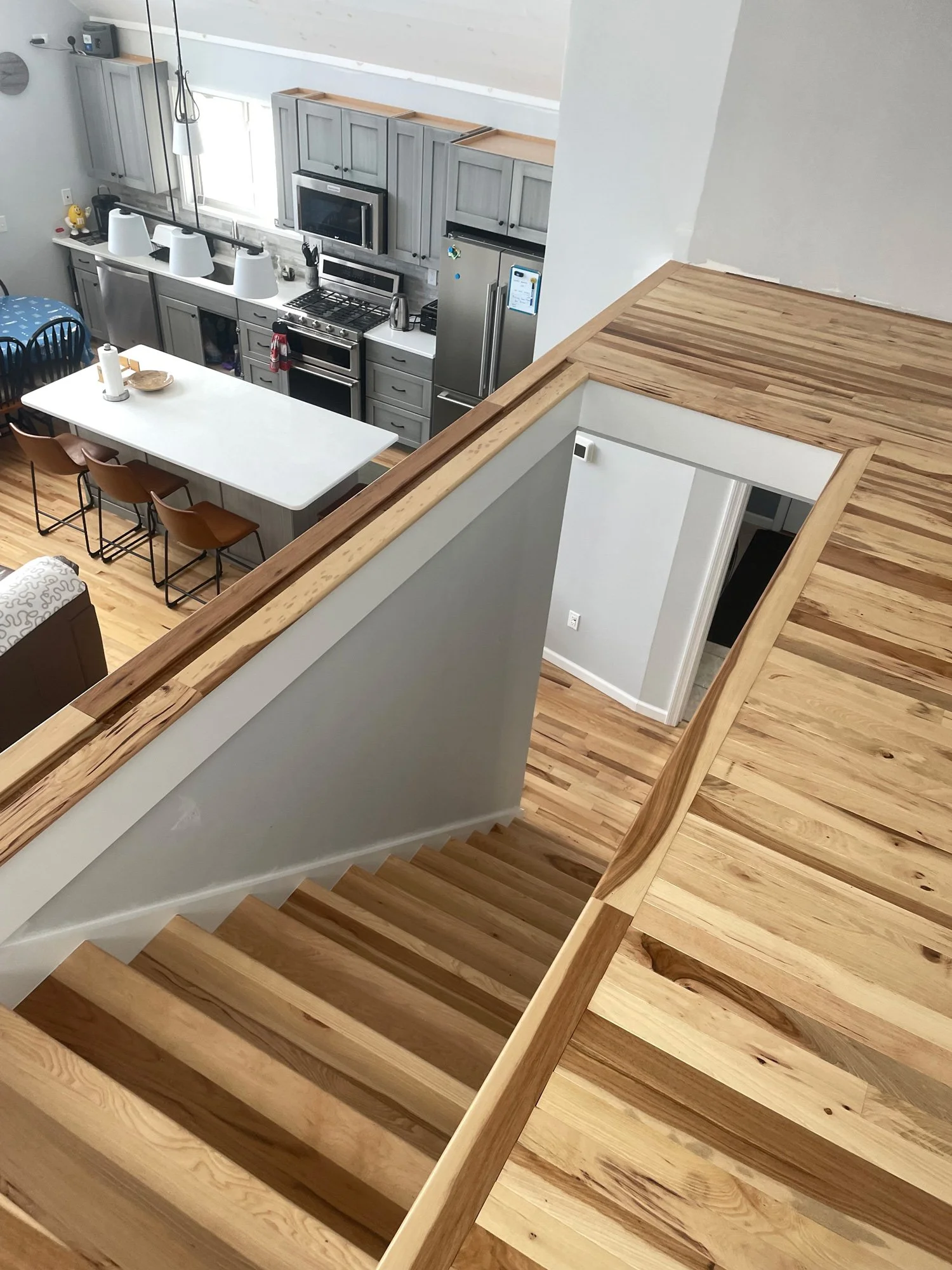 View from a staircase overlooking a modern kitchen and dining area with gray cabinets, stainless steel appliances, a white island with seating, and wooden flooring.