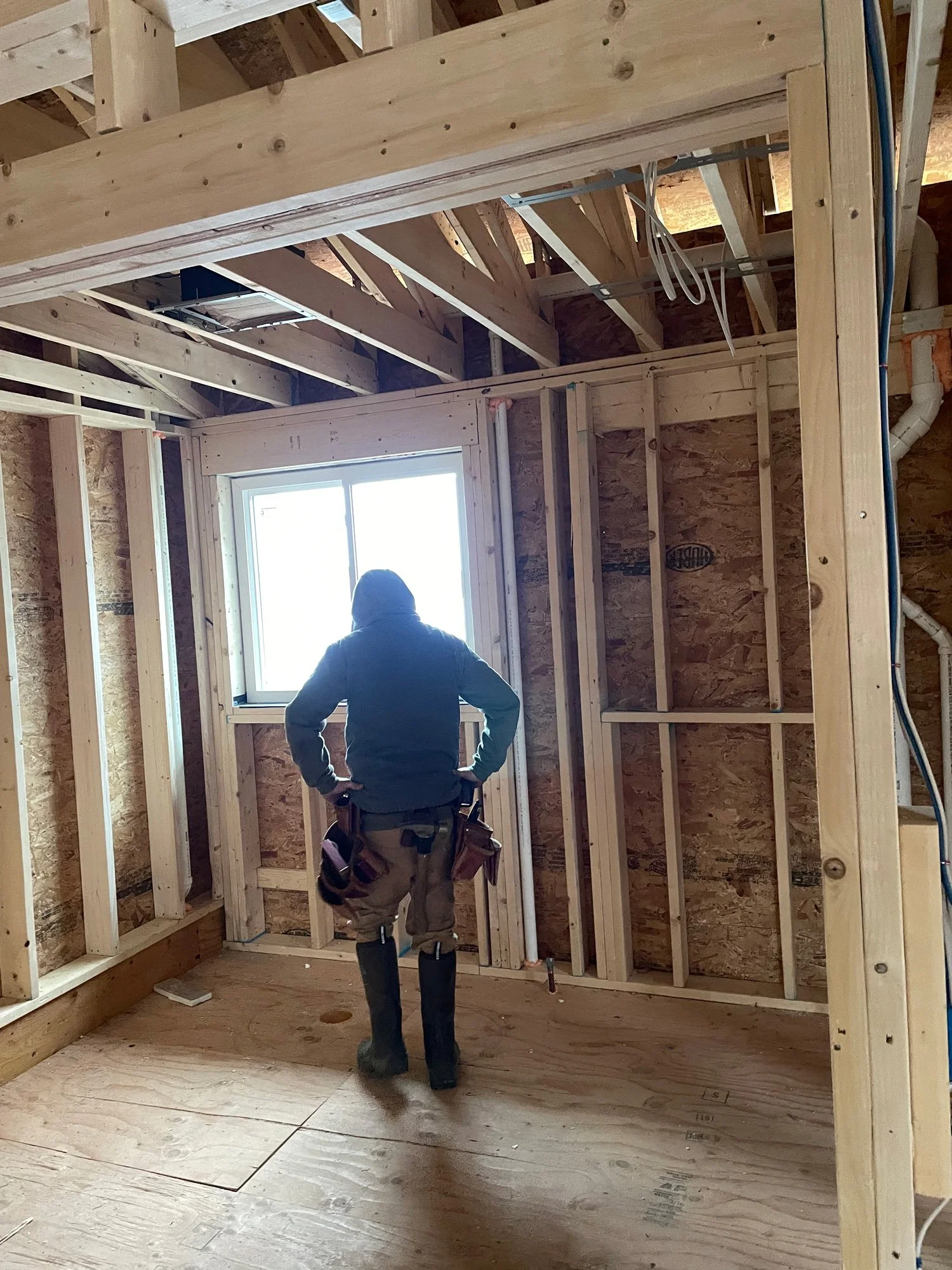 A construction worker stands inside a partially built room, looking out a window with hands on hips. The room has exposed wooden framing and plywood flooring. The worker is wearing a hoodie, work pants, and rubber boots, with a tool pouch attached to