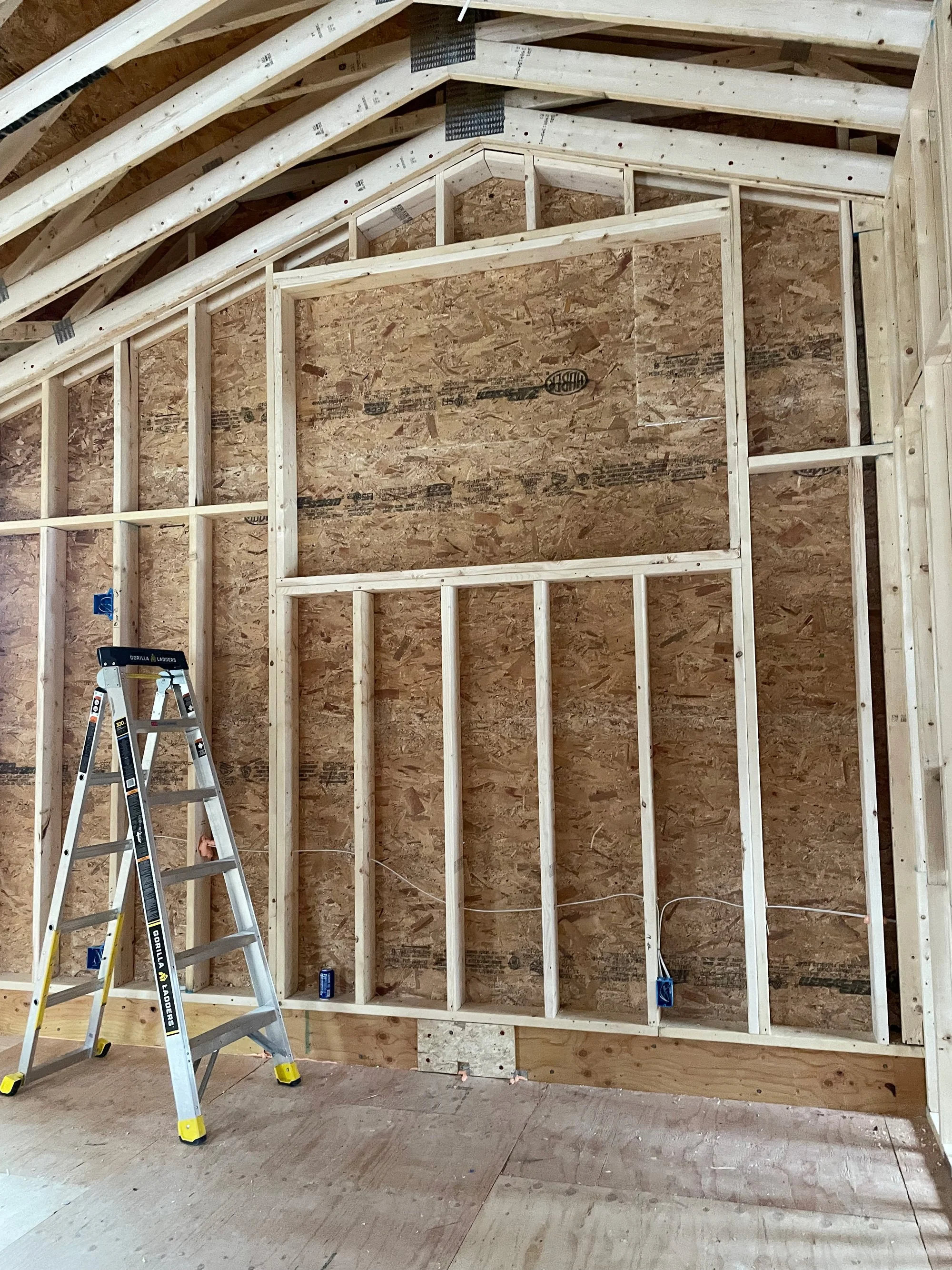 Interior of a building under construction with exposed wooden framing, a plywood wall, and a metal step ladder.