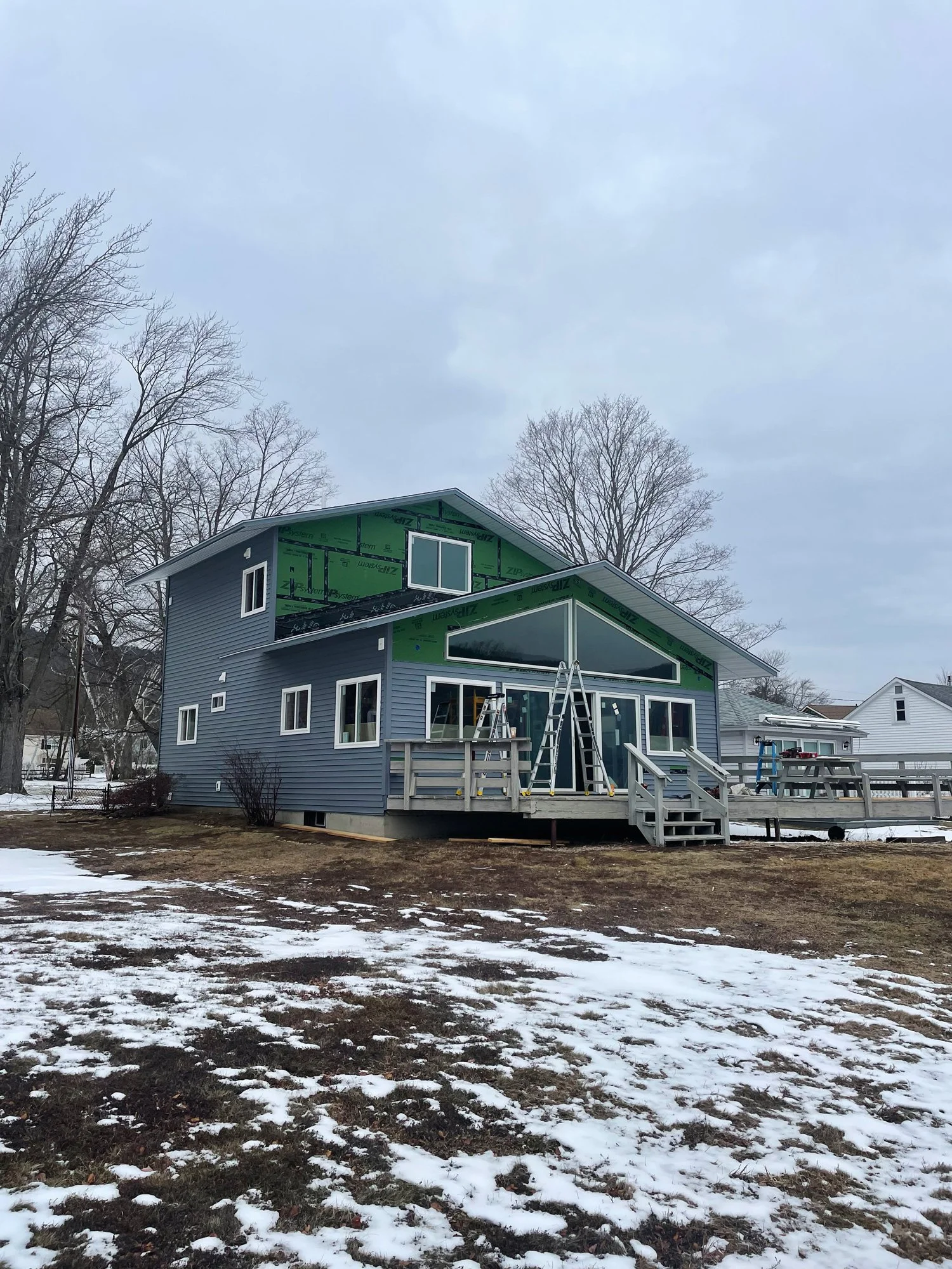 A house under construction with blue siding, multiple windows, and a large front deck with stairs, surrounded by snow and leafless trees.