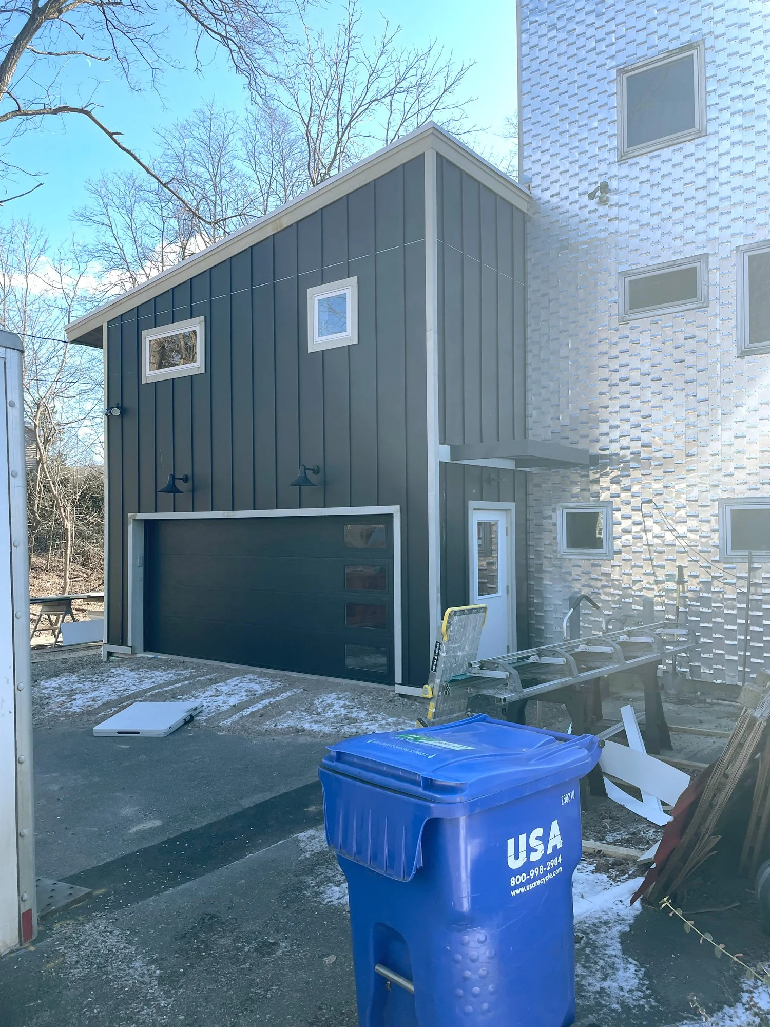 A modern two-story building under construction with dark green siding, multiple small windows, and a garage door. Construction materials and tools are visible outside, including a blue recycling bin, a ladder, and debris, with a backdrop of leafless 