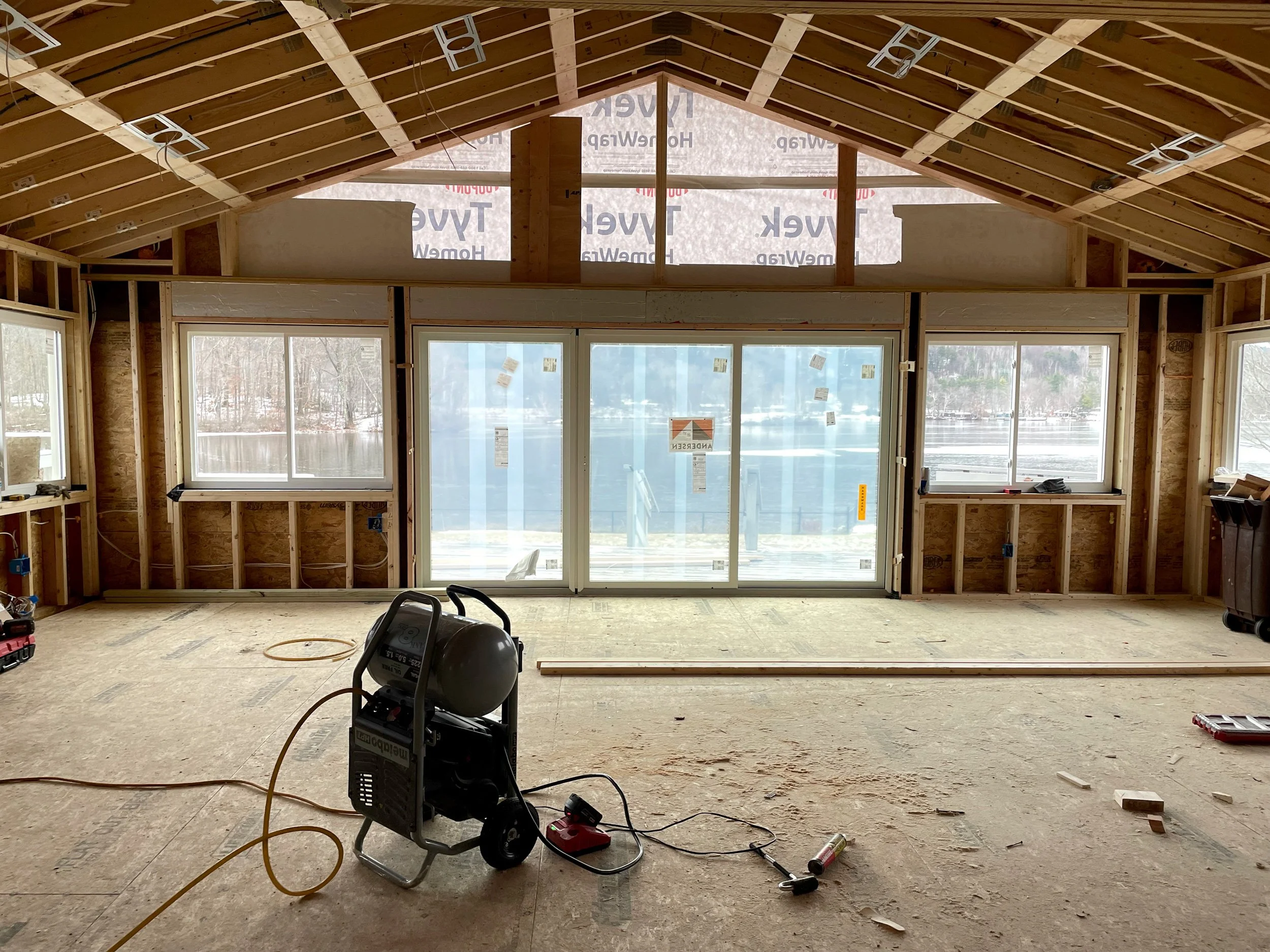 Interior view of a house under construction with exposed wooden framing, large sliding glass doors leading to a snowy lake, and construction tools on the unfinished floor.