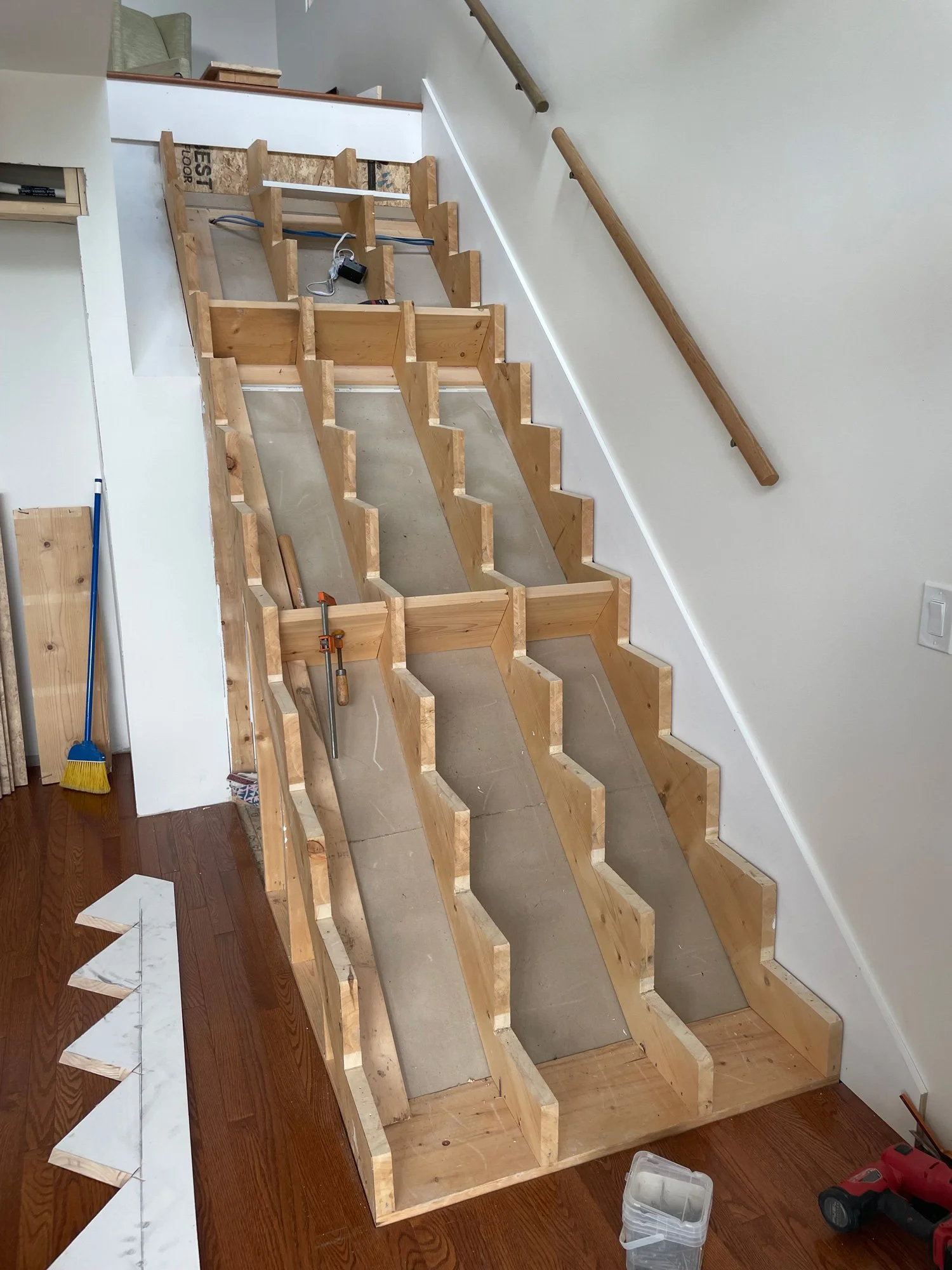 Wooden staircase frame under construction with multiple brackets and supports, a handrail attached on the wall, and construction tools nearby.