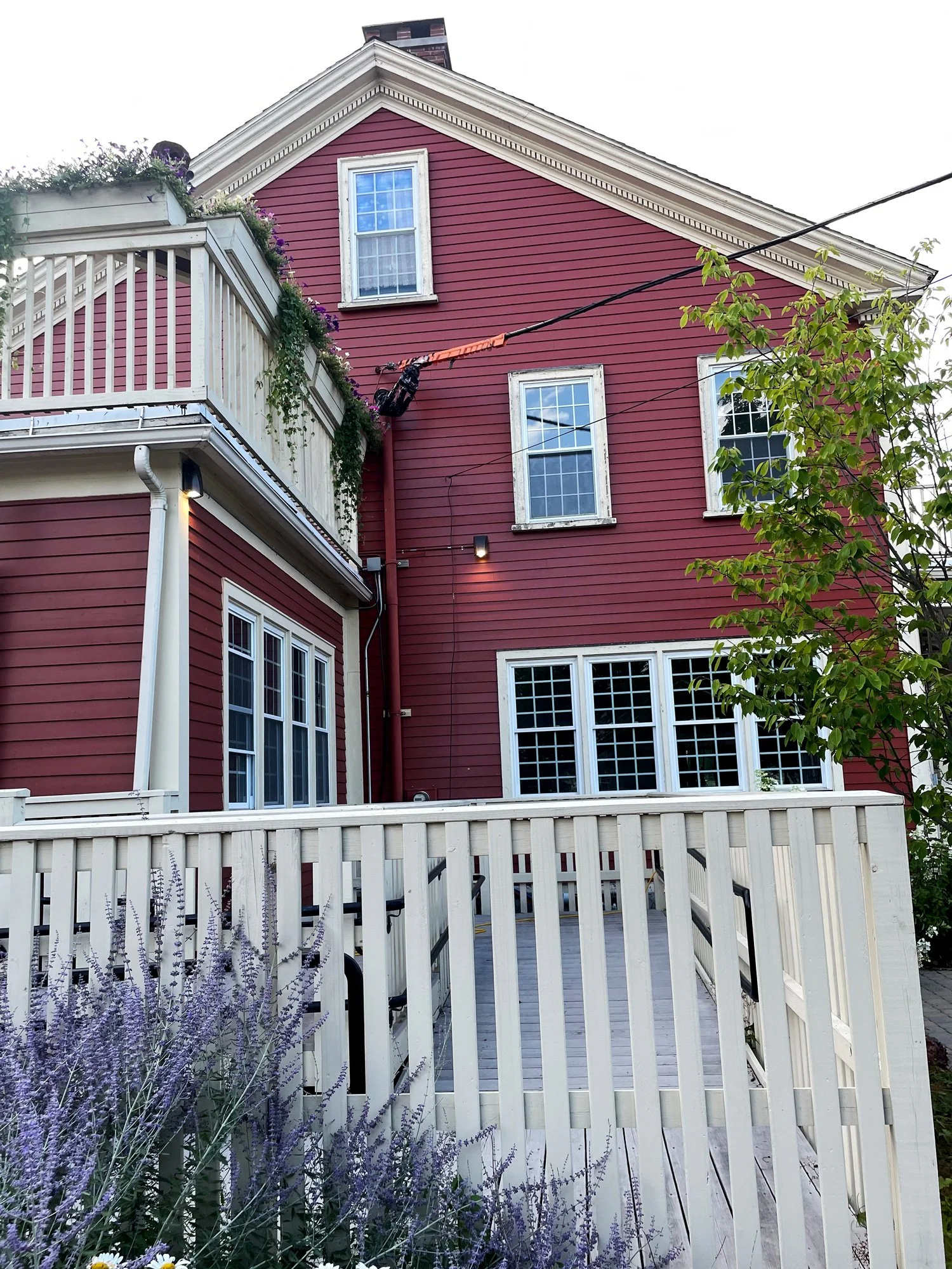 Red wooden house with white trim, three prominent windows, a balcony with purple flowers, a tree on the right, and a wooden deck with white railing in the foreground.