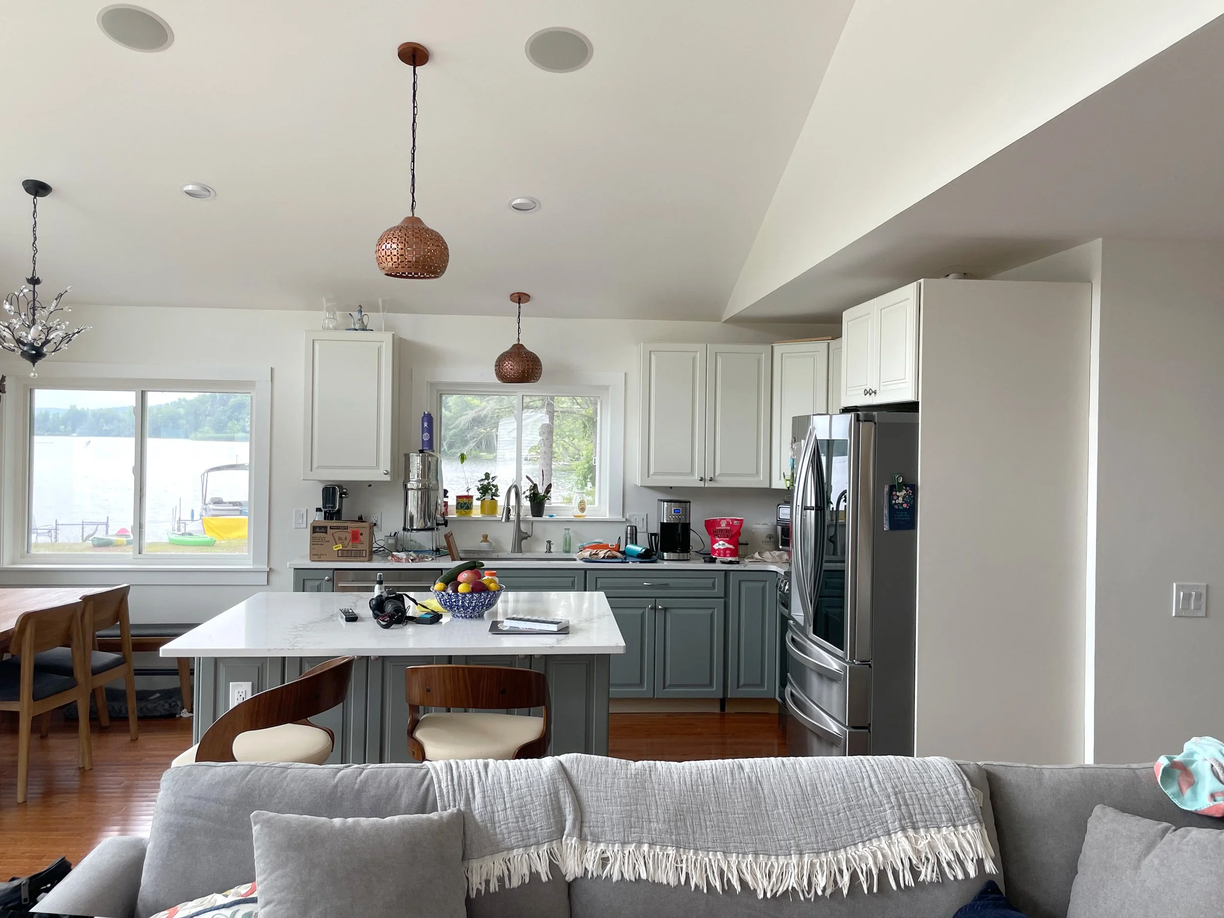 Kitchen with white cabinets, a marble island, and large windows overlooking a lake. The island has a bowl of fruit and remote control on it. There are three hanging pendant lights with woven covers. Part of a grey couch with a fringe blanket is visib