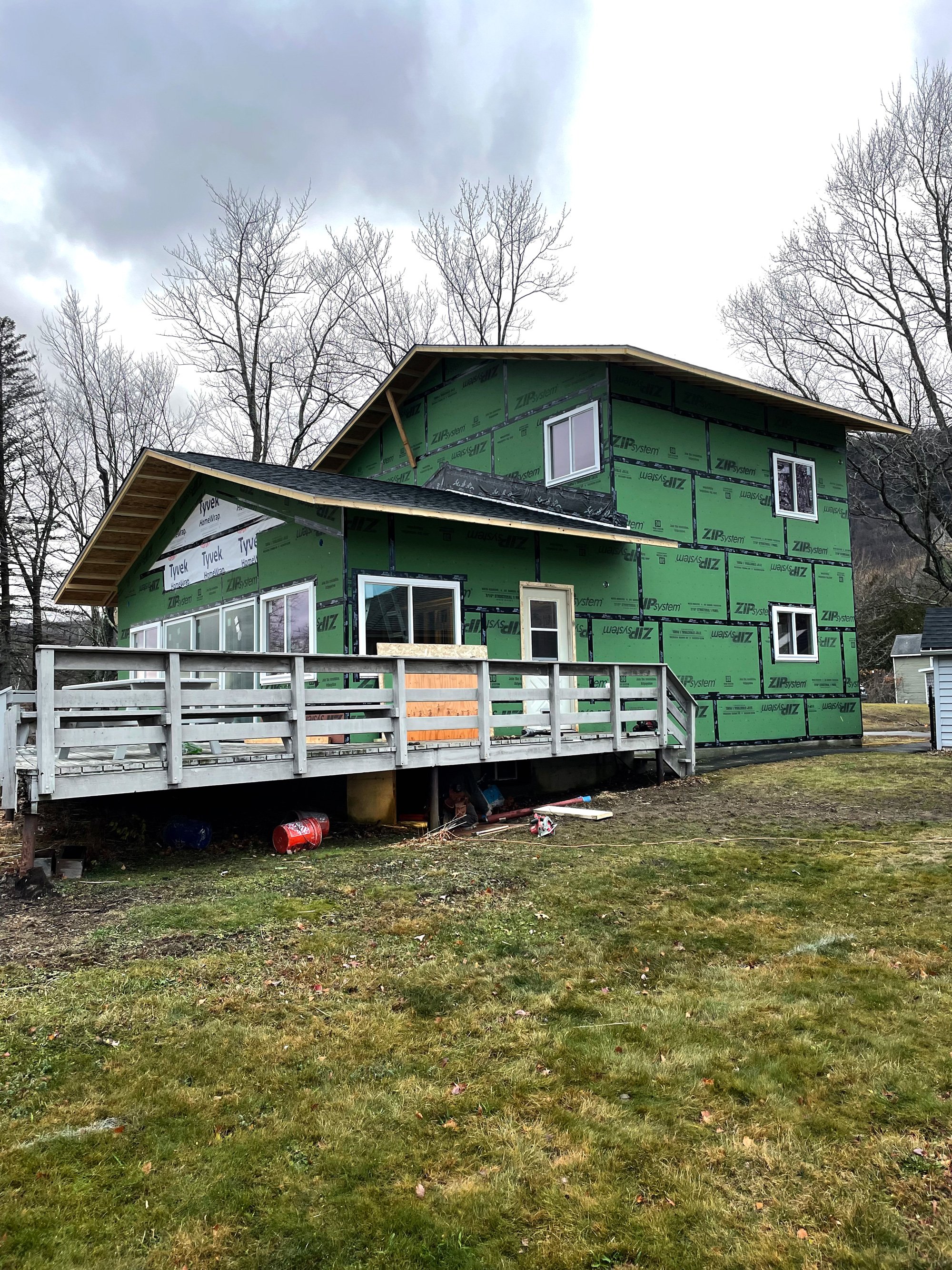 Photo of a house under construction with green house wrap, multiple windows, a deck, and a sloped roof, surrounded by leafless trees and a cloudy sky.