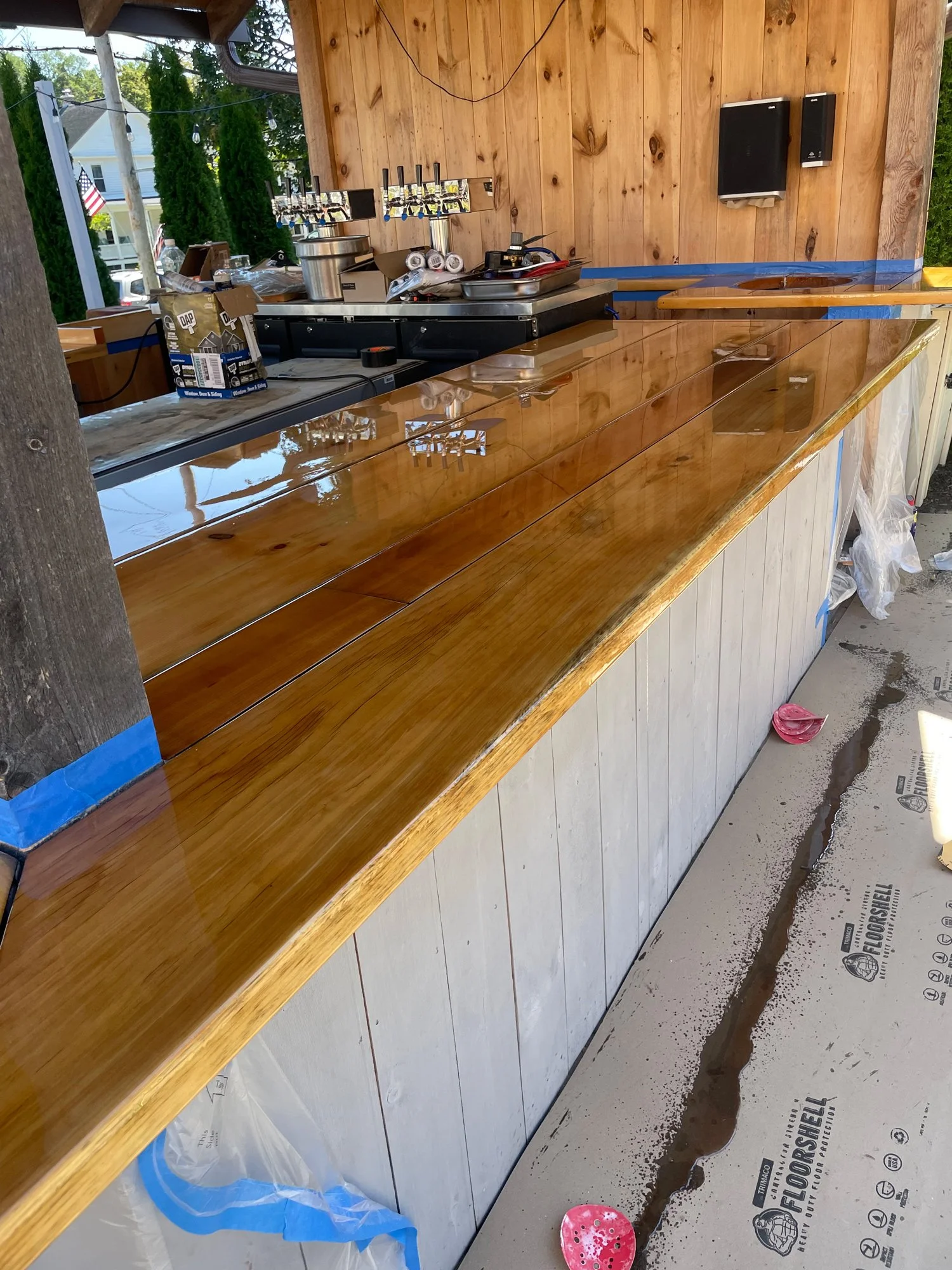 A wooden bar counter under construction with a glossy finish, next to a plywood wall, and a deck area with construction materials and tools visible. In the background, there are trees and houses through an open outdoor space.