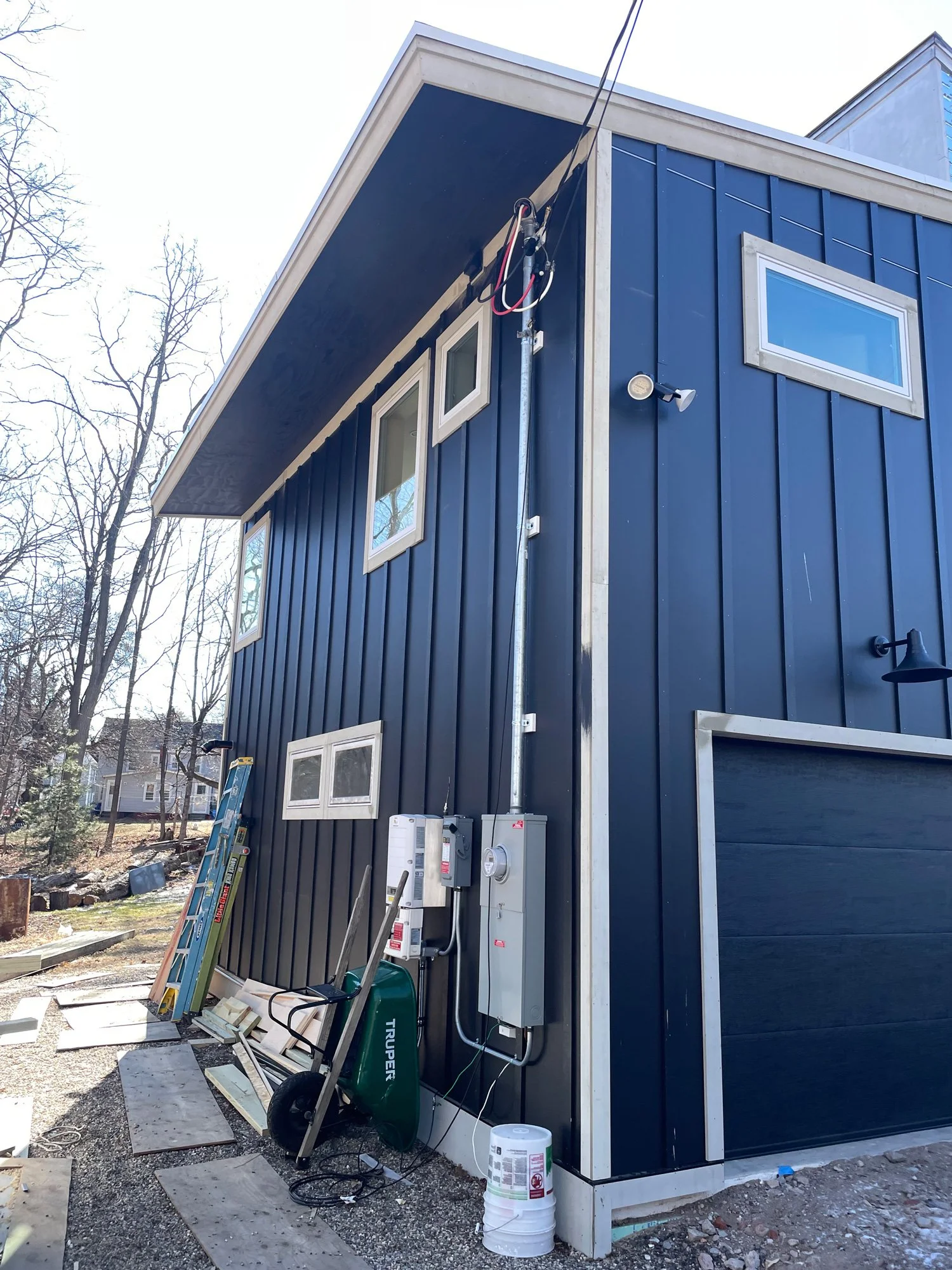 Back of a modern blue house under construction with tools and supplies on the ground, electrical panels on the wall, and a wheelbarrow.