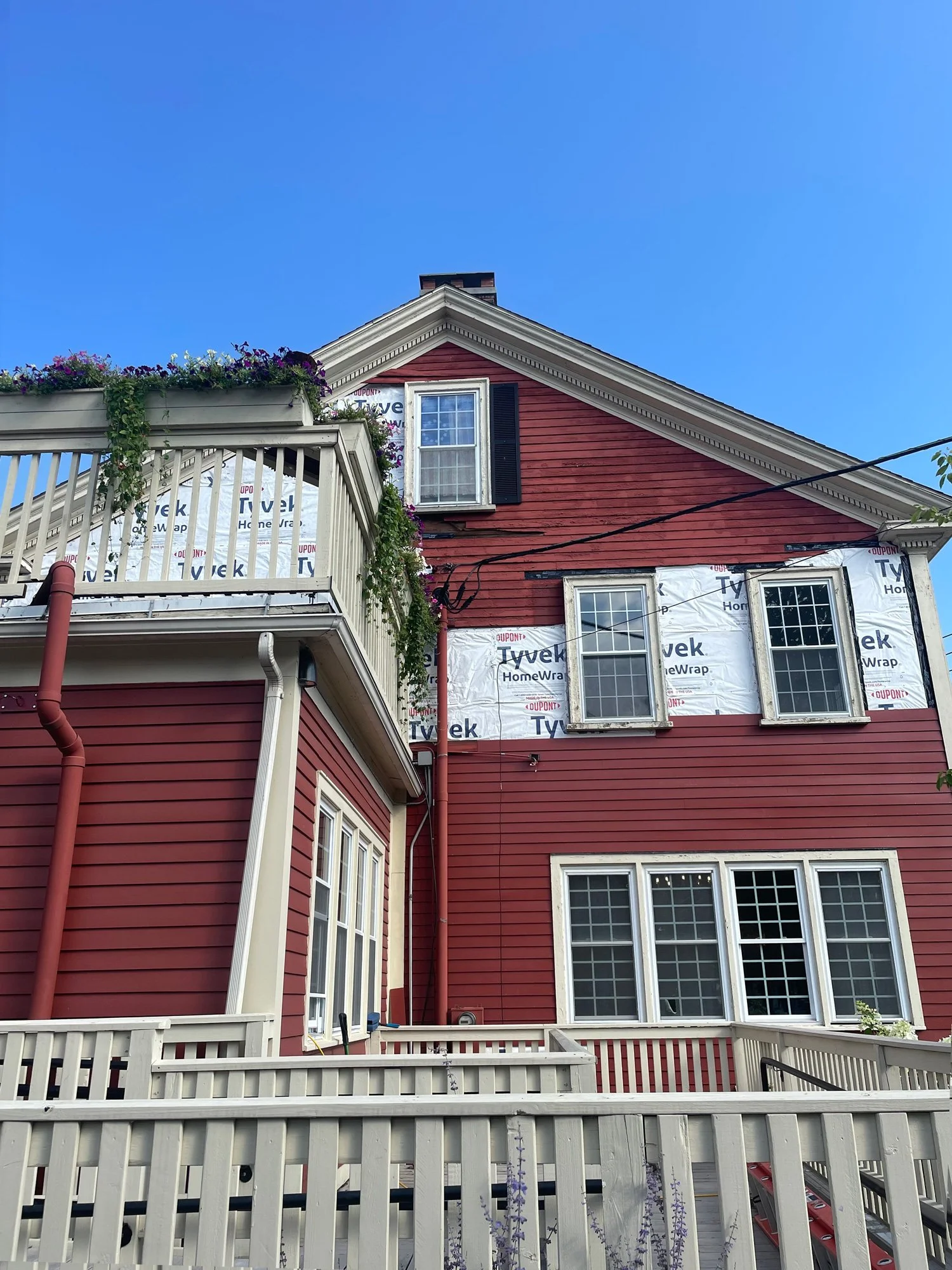The house is under renovation with visible Tyvek HomeWrap on the upper part of the house, red exterior siding, multiple windows, a balcony with flower boxes, and a white fence in the foreground.