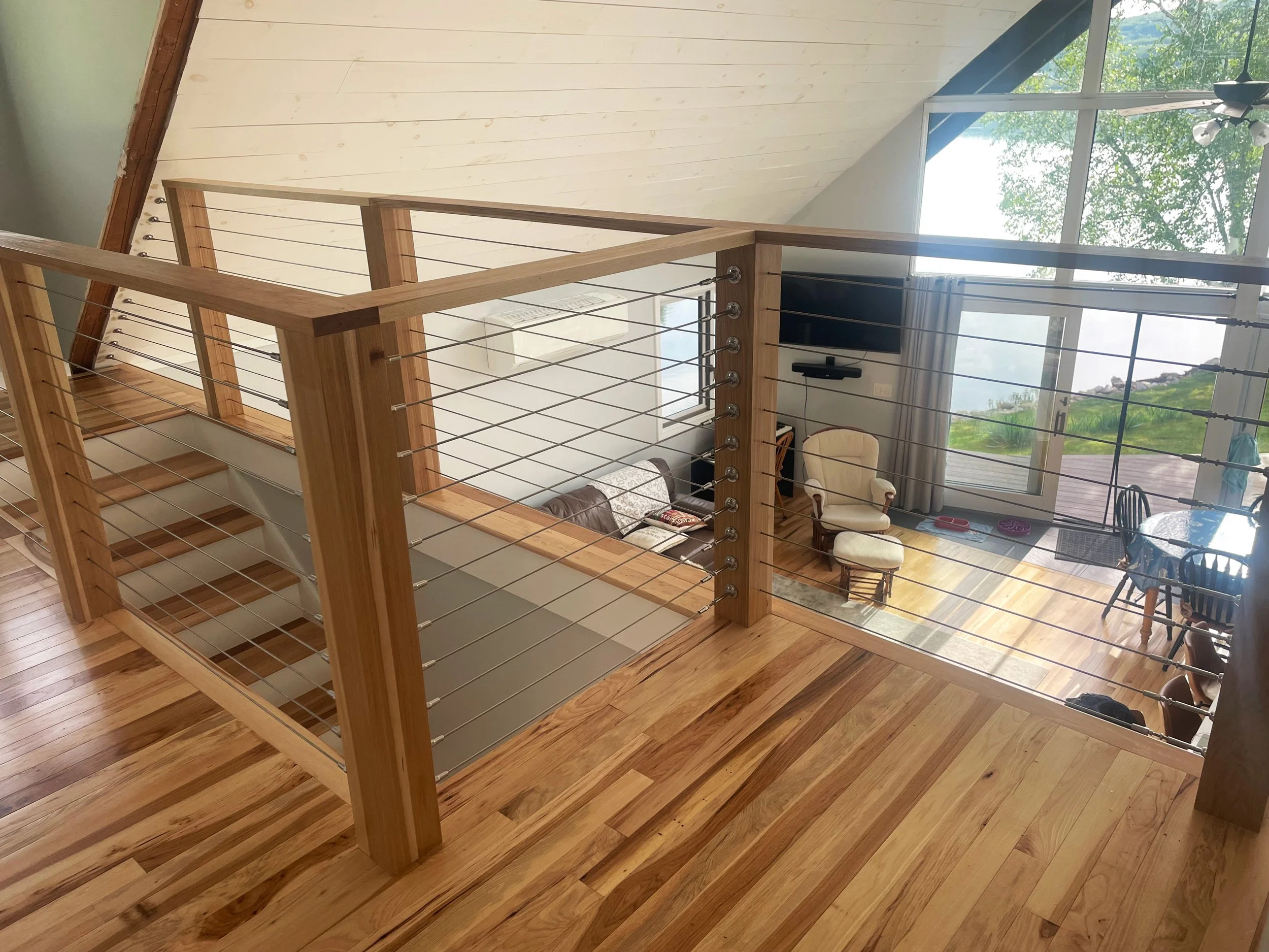 View of a loft area overlooking a living room with large windows showing an outdoor landscape. The loft has a wooden floor and a modern cable railing.