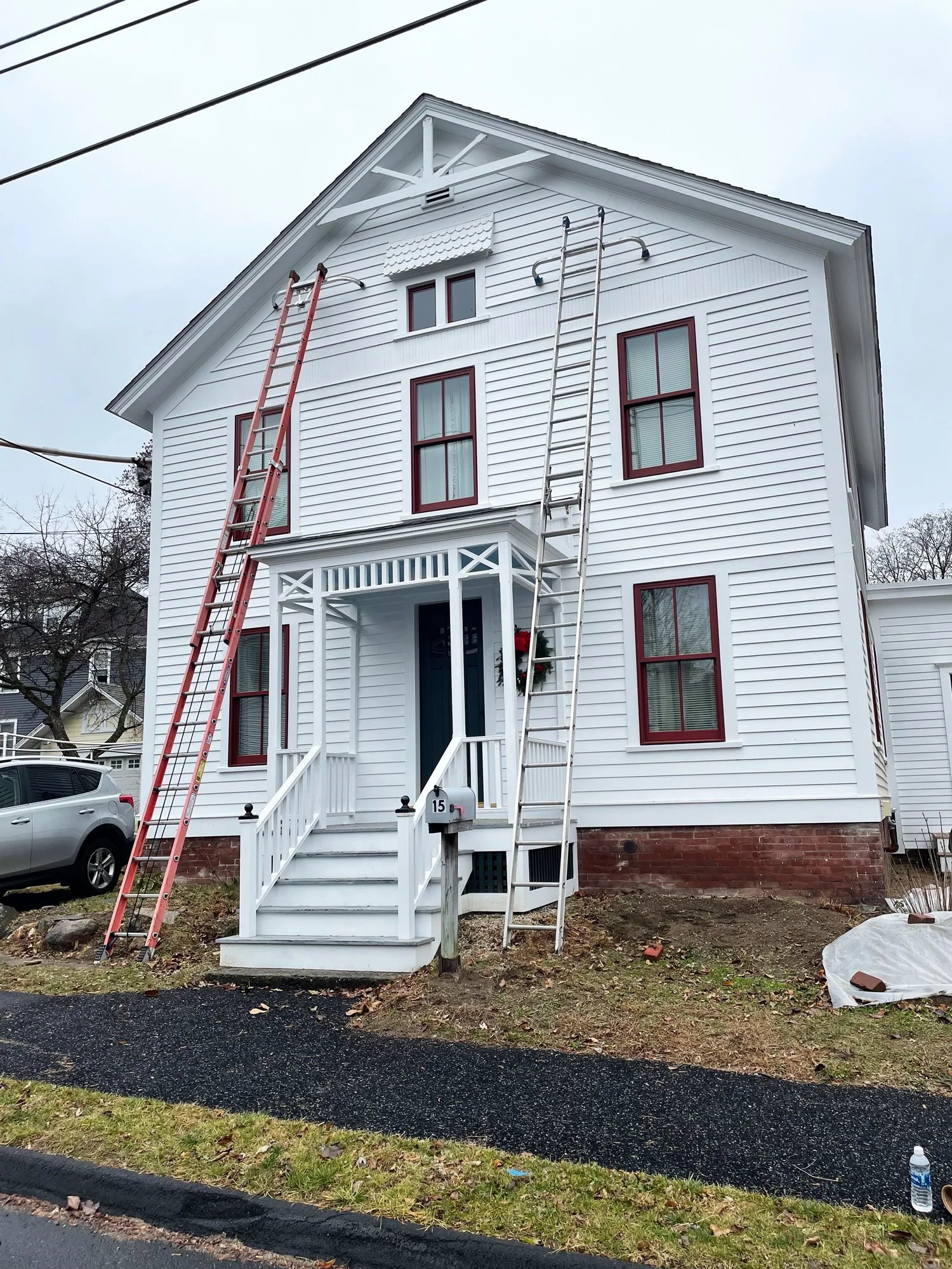 A white two-story house with red window frames, front porch, and two long ladders leaning against the house, one red and one silver, indicating ongoing work or renovation.