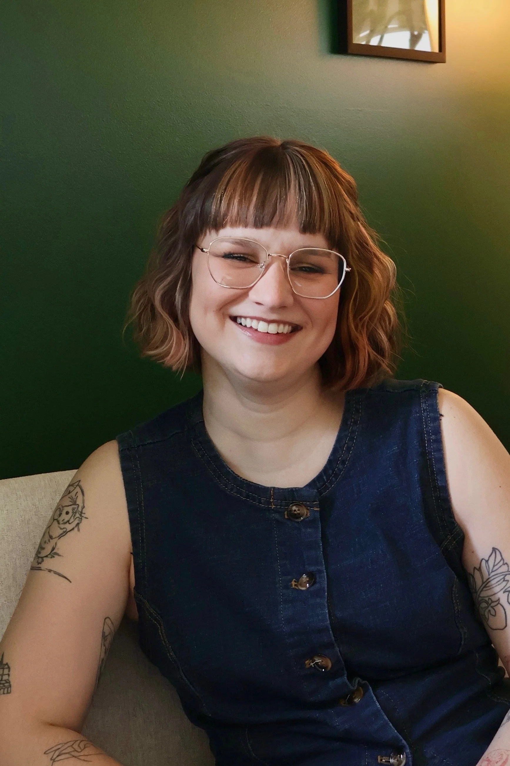 A smiling woman with shoulder-length light brown hair, wearing glasses and a light-colored striped shirt, sitting on a beige sofa with pillows in the background.
