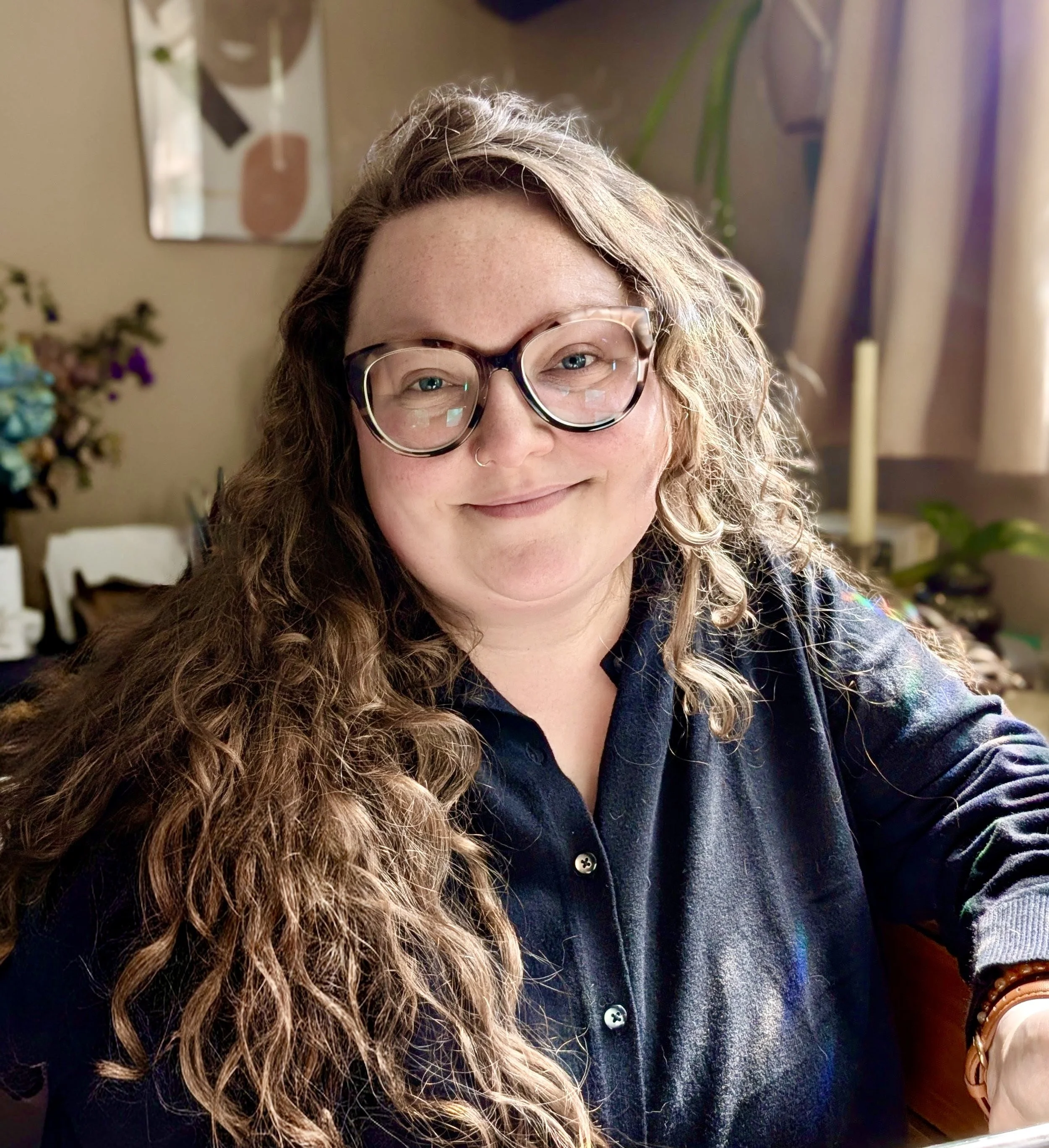 Close-up of a smiling woman with curly brown hair, wearing large tortoiseshell glasses and a denim shirt.
