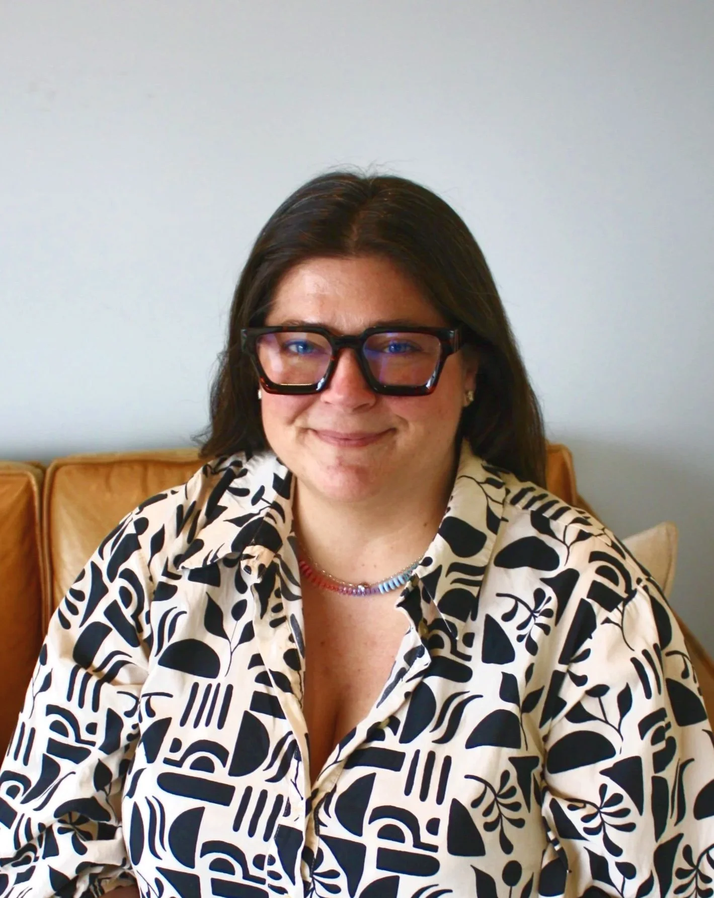 A woman with shoulder-length dark hair wearing large black glasses and a black-and-white patterned dress, sitting on a tan couch with a light-colored wall in the background.