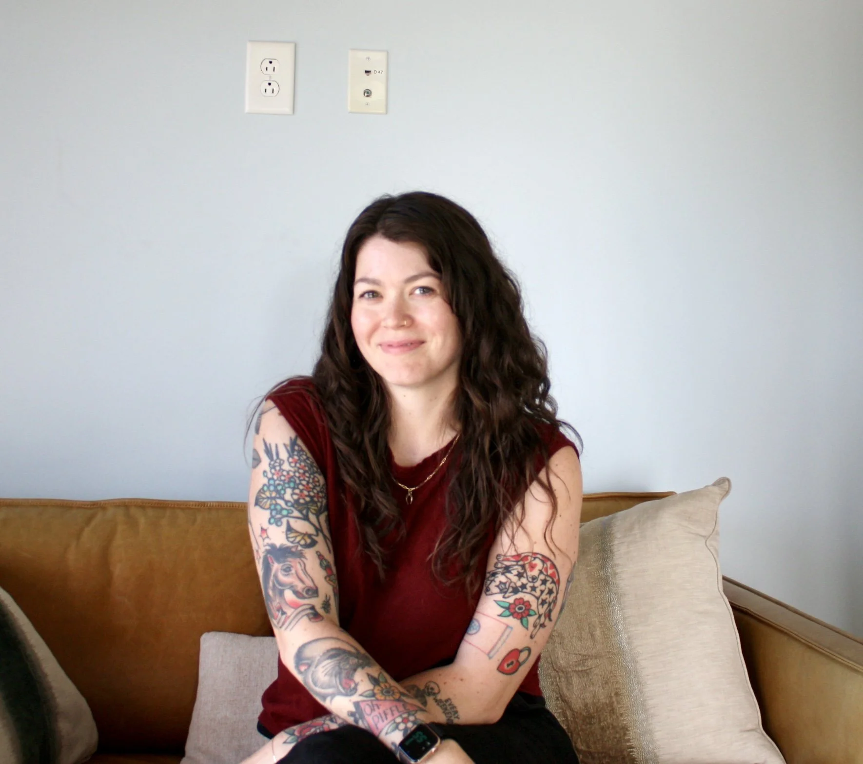 A young woman with long wavy brown hair, tattoos on her arms, and a nose ring, smiling while sitting on a blue chair in a room with beige walls.