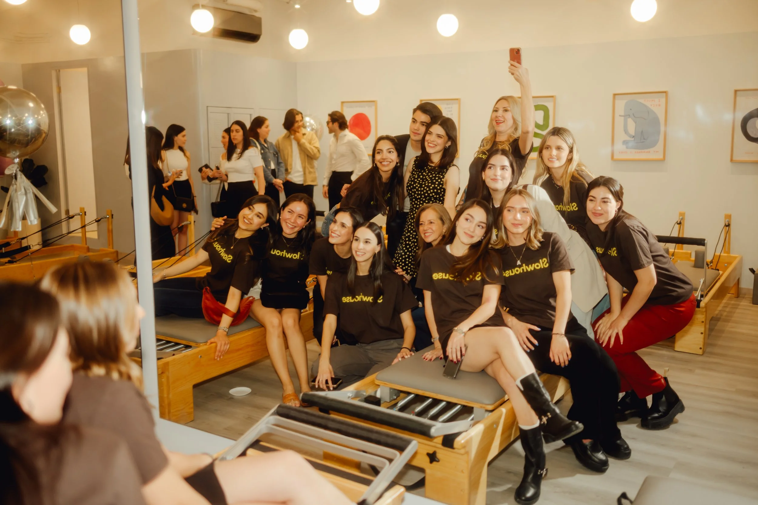 Grupo de mujeres y algunas personas en el fondo tomando una selfie en un ambiente interior con iluminación cálida y decoraciones modernas.