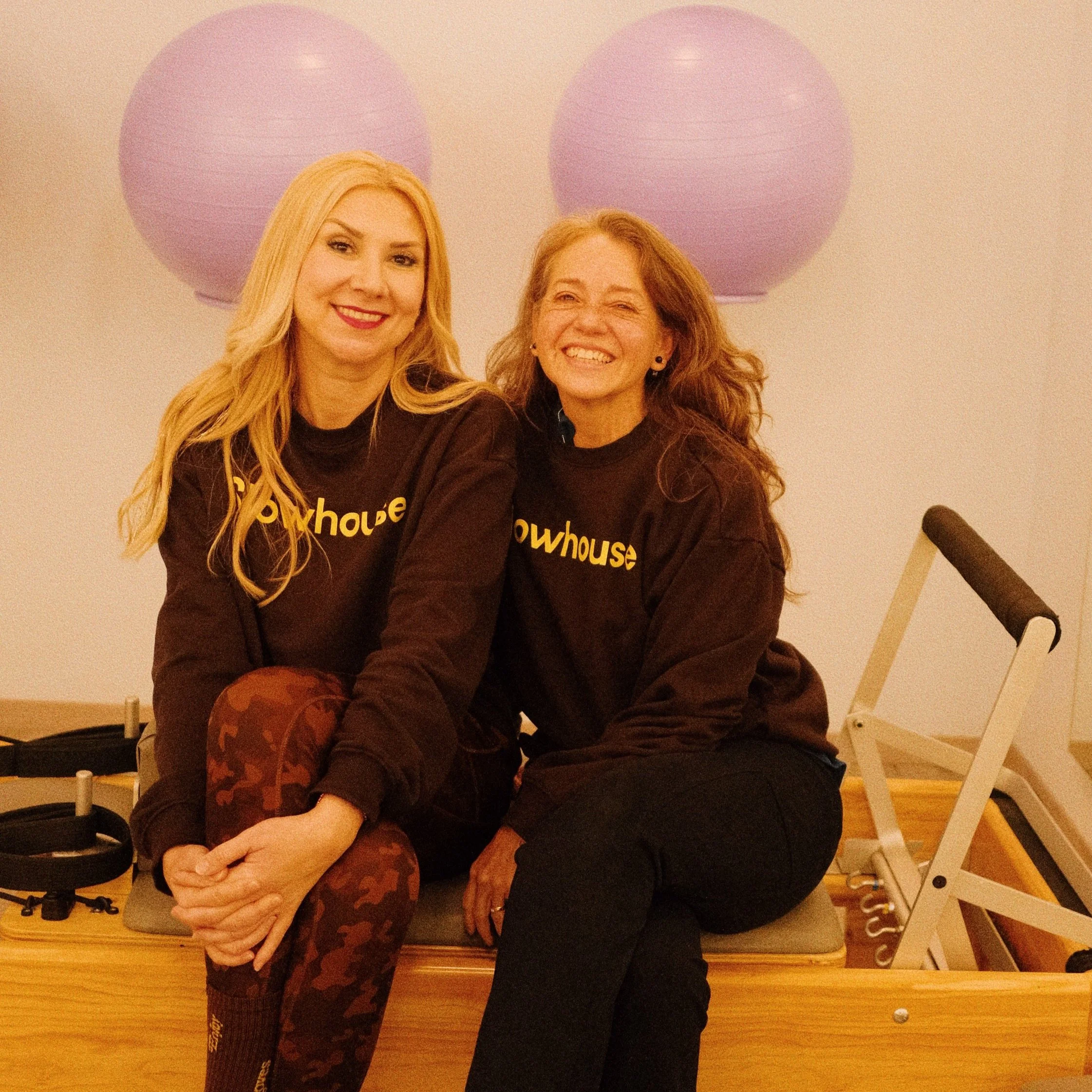 Dos mujeres con camisetas negras que dicen 'Showhouse', sentadas en un equipo de pilates en un entorno con lámparas de papel moradas en la pared.