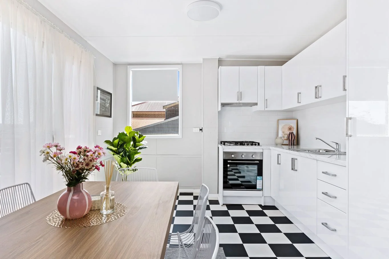 Modern white kitchen with checkered black and white floor, dining table with pink flowers in a vase, natural light from window, and minimalist decor.