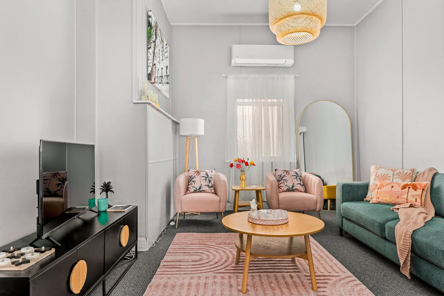 Living room with pastel pink armchairs, a green sofa, a wooden coffee table, a pink patterned rug, a large mirror, a TV on a black stand, and decorative items including a vase with red and yellow flowers, throw pillows, and a floor lamp.