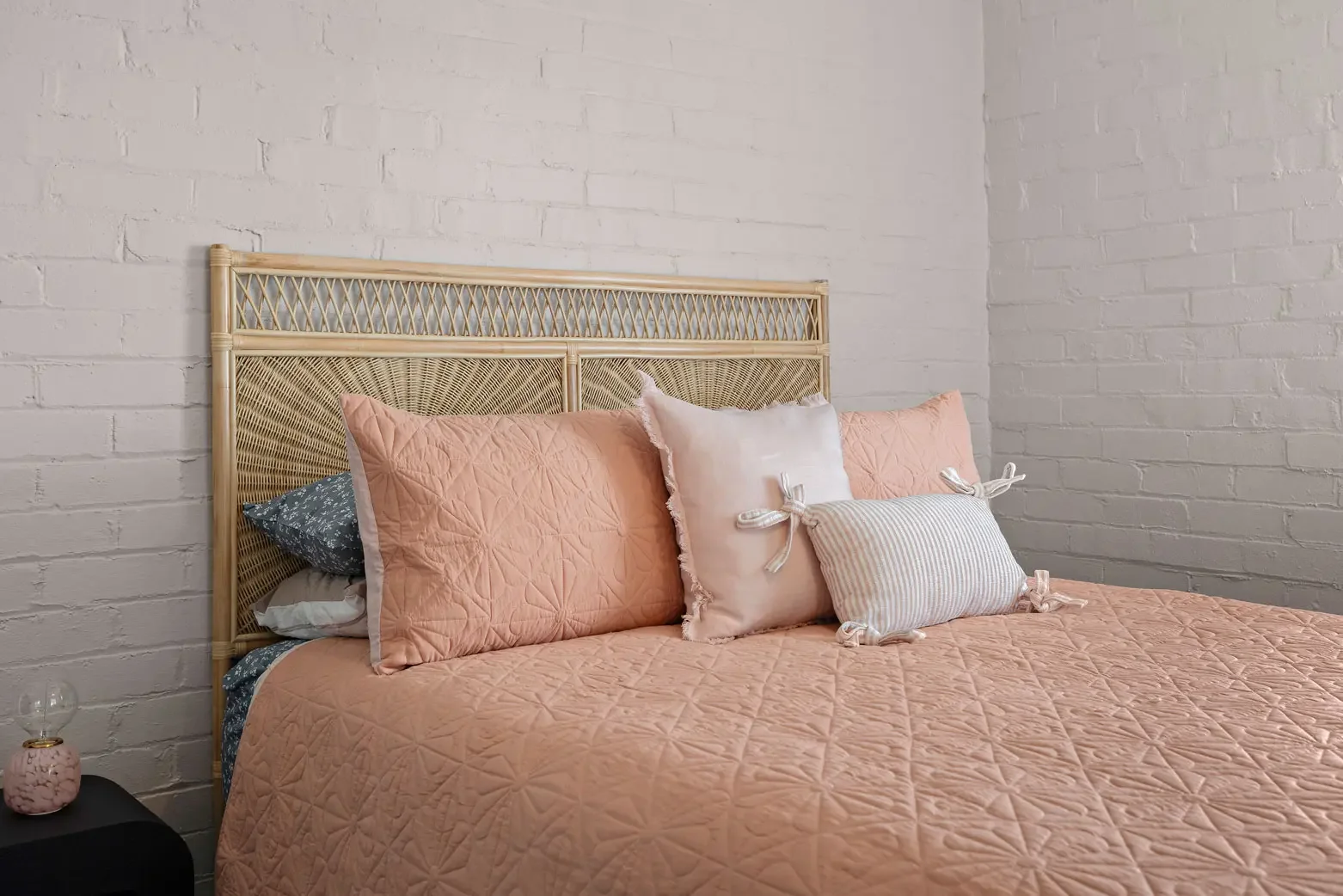 A bedroom with a bed covered in a pink quilt and decorated with pink, white, and striped pillows, and a rattan headboard against a white brick wall.