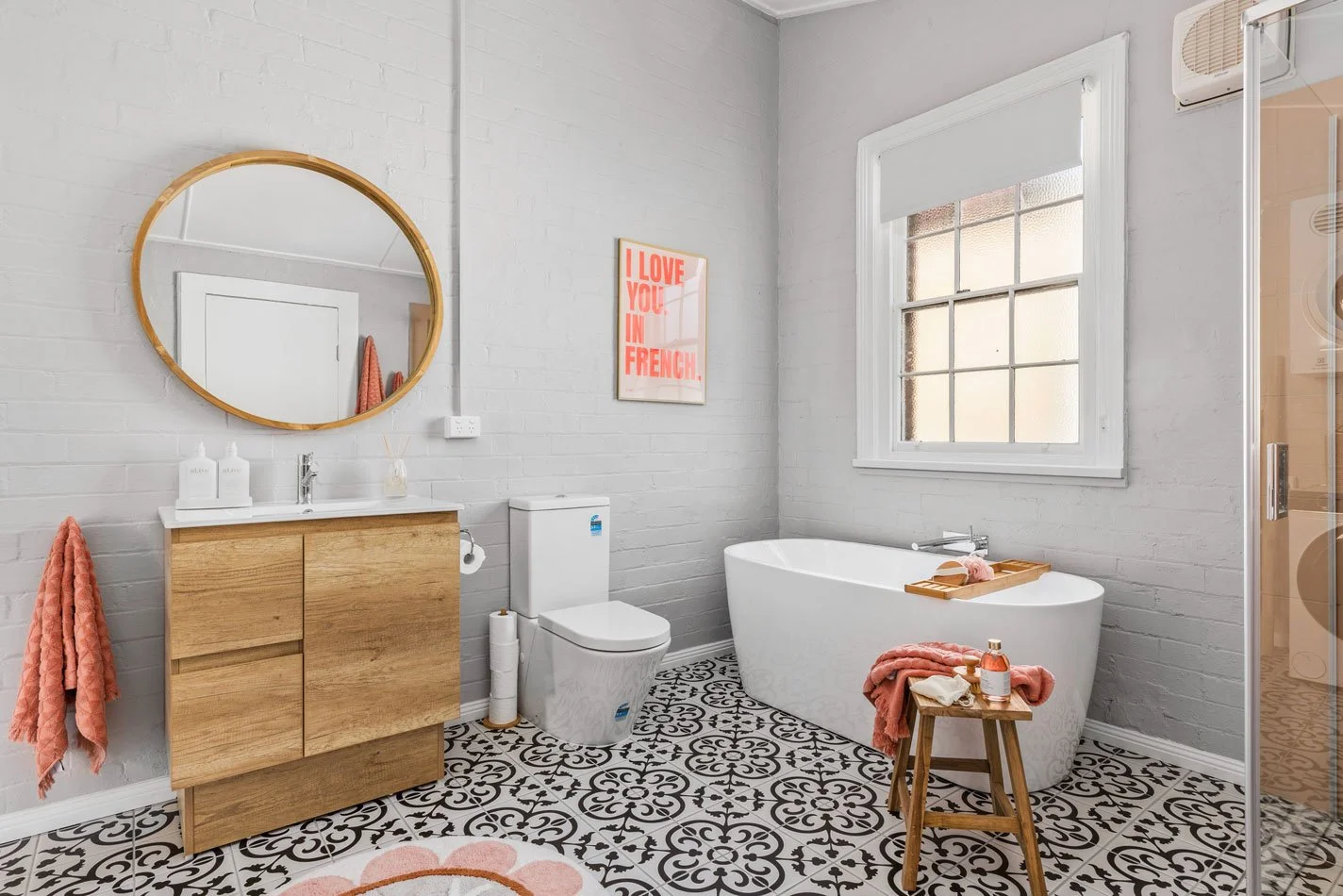 Modern bathroom with white brick walls, a wooden vanity with a round mirror, a white bathtub, a toilet, a window, and pink accessories like towels and soap.