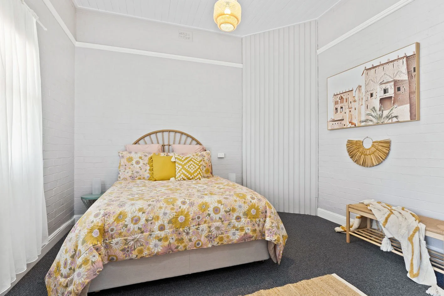 A bedroom with a bed covered in a floral yellow, pink, and white bedspread, four pillows including two pink, one yellow, and a patterned yellow and white pillow. The room has white brick walls, a white ceiling, and a window with sheer white curtains. There is a teal side table with candles, artwork on the wall, and a wooden bench with a blanket on it.