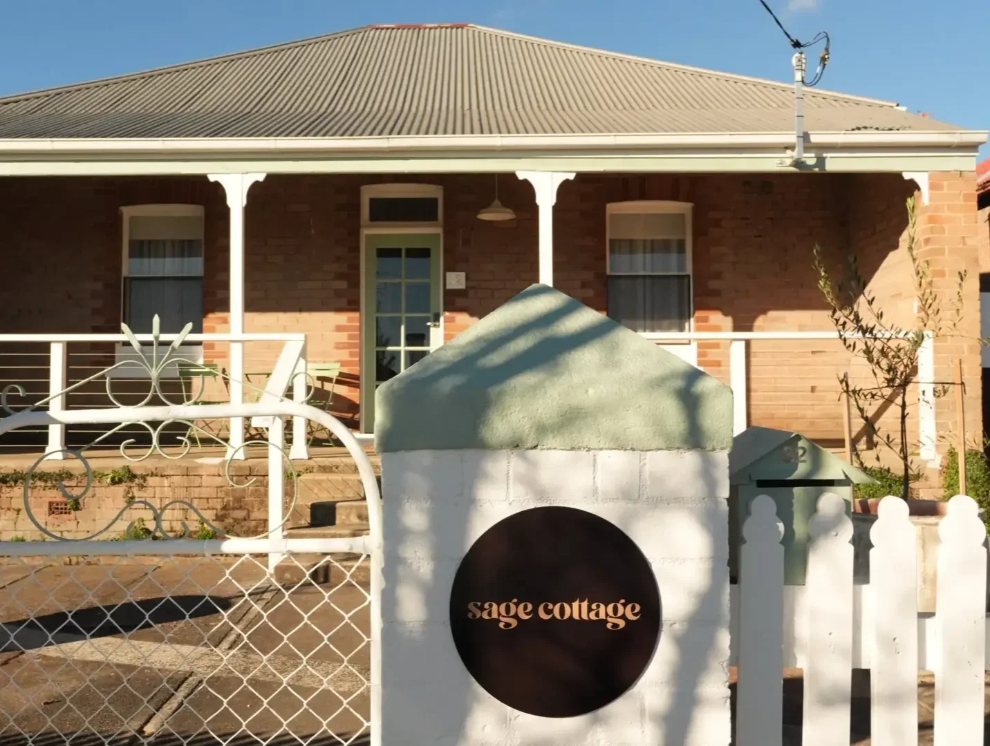 Front view of a brick house with a porch, white railing, and a sign that reads 'sage cottage' near the gate. The house has a metal roof, a door, and two windows. There is a white gate and front yard with plants and shadows from nearby trees.
