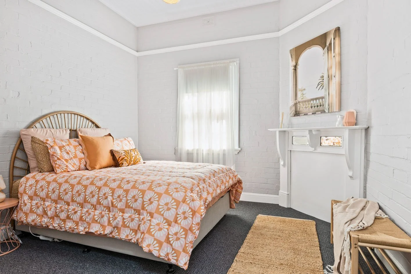 Bedroom with white brick walls, a window with white curtains, and a bed with a geometric orange and white floral bedspread and multiple pillows. A mirror above a white mantle, a small beige rug, and a bench with a beige throw blanket are also visible.
