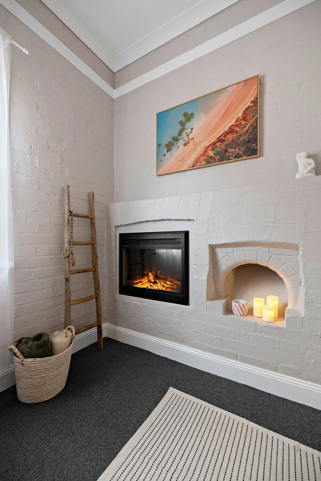 A cozy living room corner with a fireplace, a modern framed artwork above, a ladder leaning against the wall with a basket of blankets, and a small nook with candles.