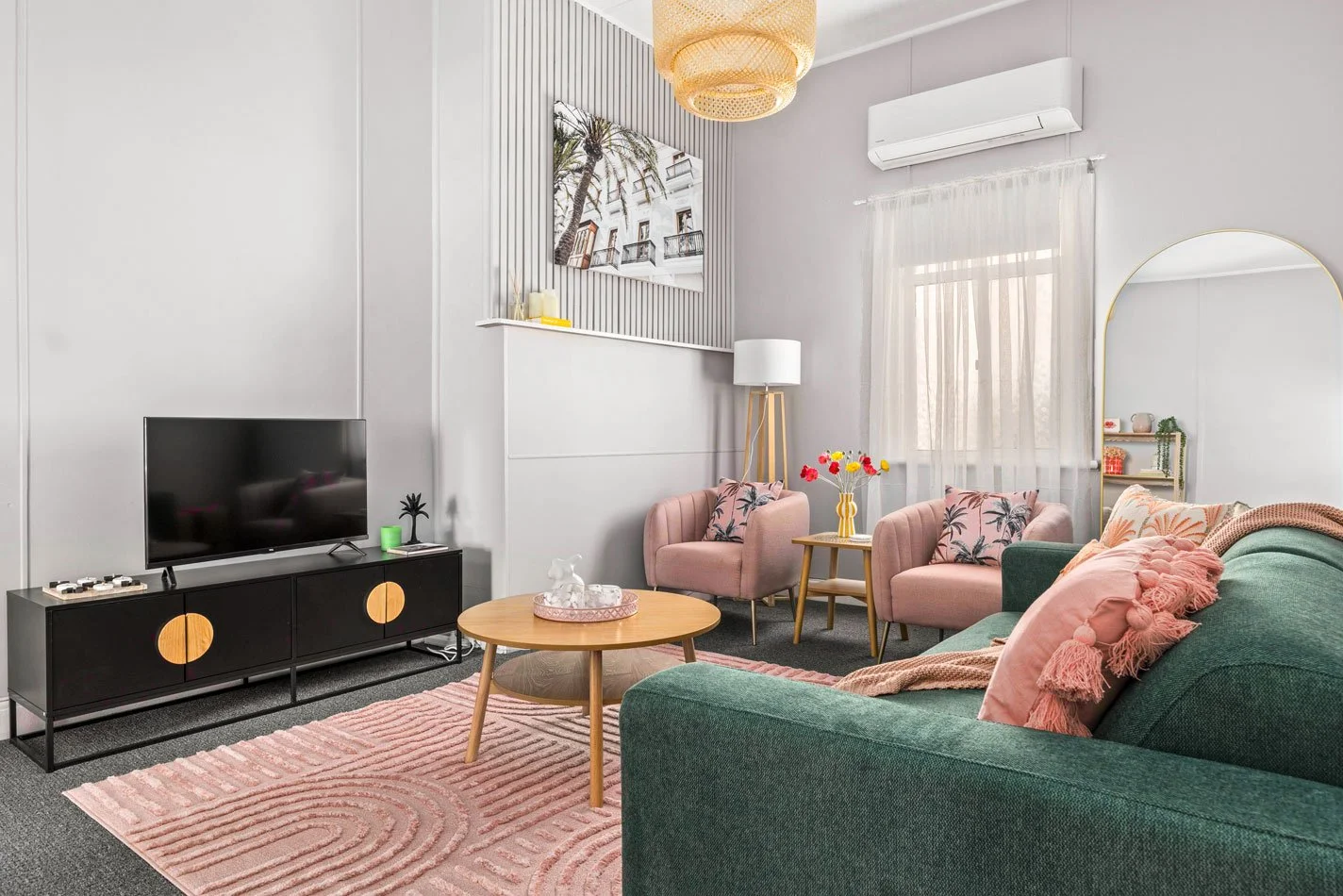 A brightly decorated living room with a green sofa, pink armchairs, a wooden coffee table, a pink rug, a wall-mounted TV, and a window with white curtains.