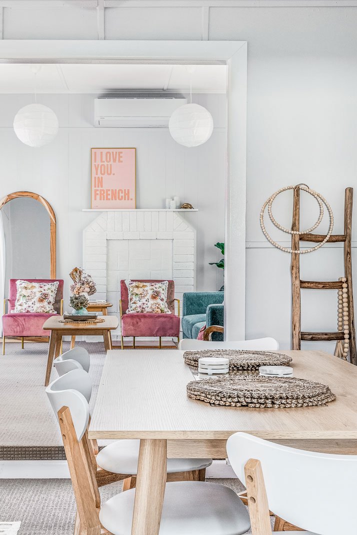 Bright and airy living and dining area with a wooden dining table and white chairs, pink and green armchairs, decorative pillows, a fireplace, wall art that says 'I love you in French,' hanging paper lanterns, and a wooden ladder with beaded decor.