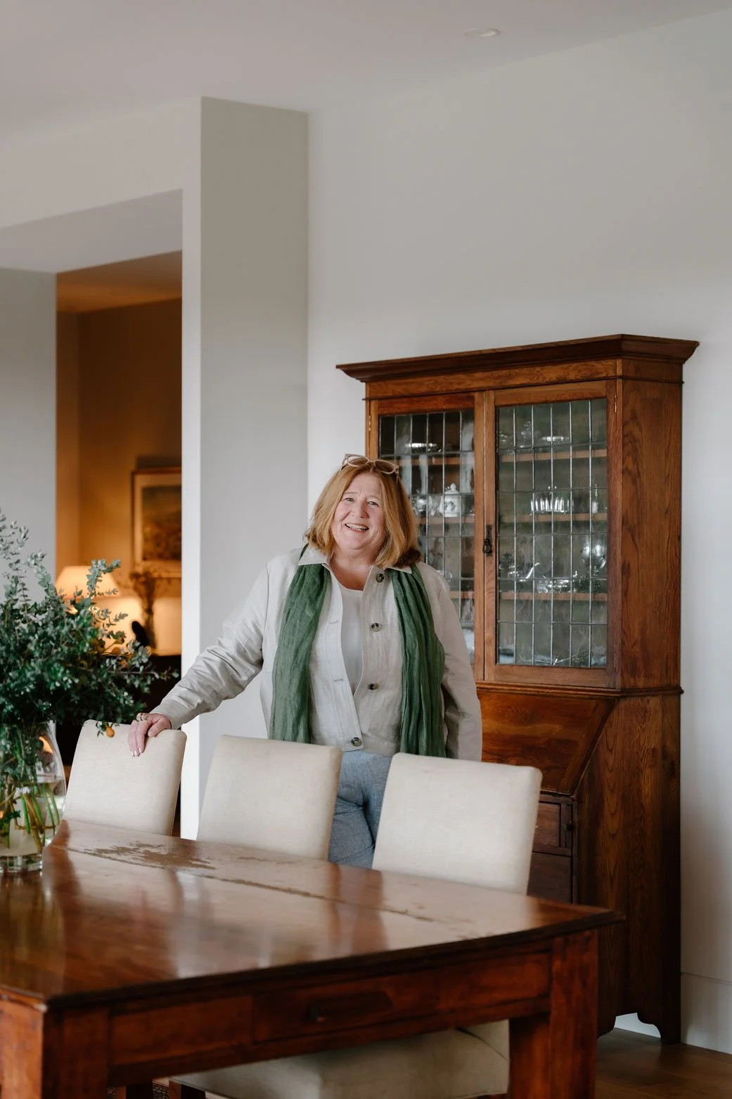 Suzie standing behind a wooden dining table with cream-coloured chairs, smiling, in a well-lit dining room with a wooden china cabinet behind her.