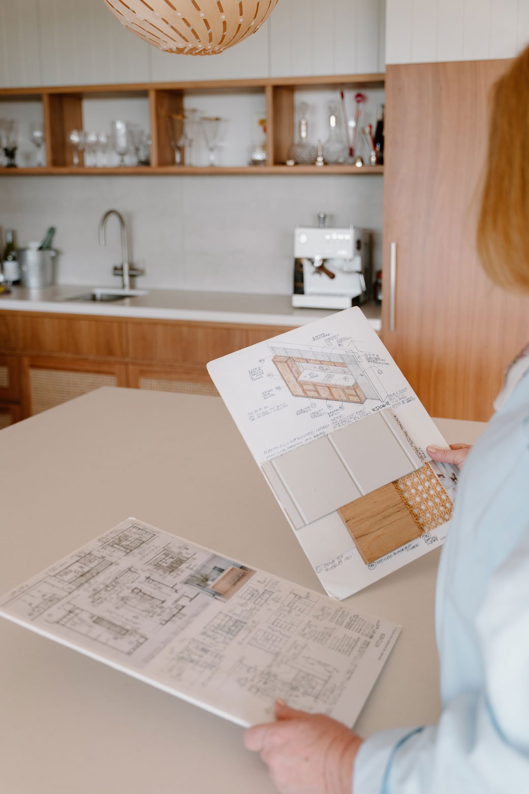 Suzie holding design moodboard in a modern kitchen with wooden cabinets and open shelves.