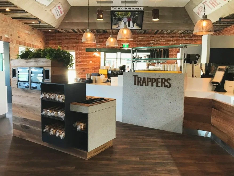Coffee shop counter at Trappers cafe. There are hanging lights, shelves with cups and bottles, and a brick wall background. A snack display with packaged snacks is in the foreground.