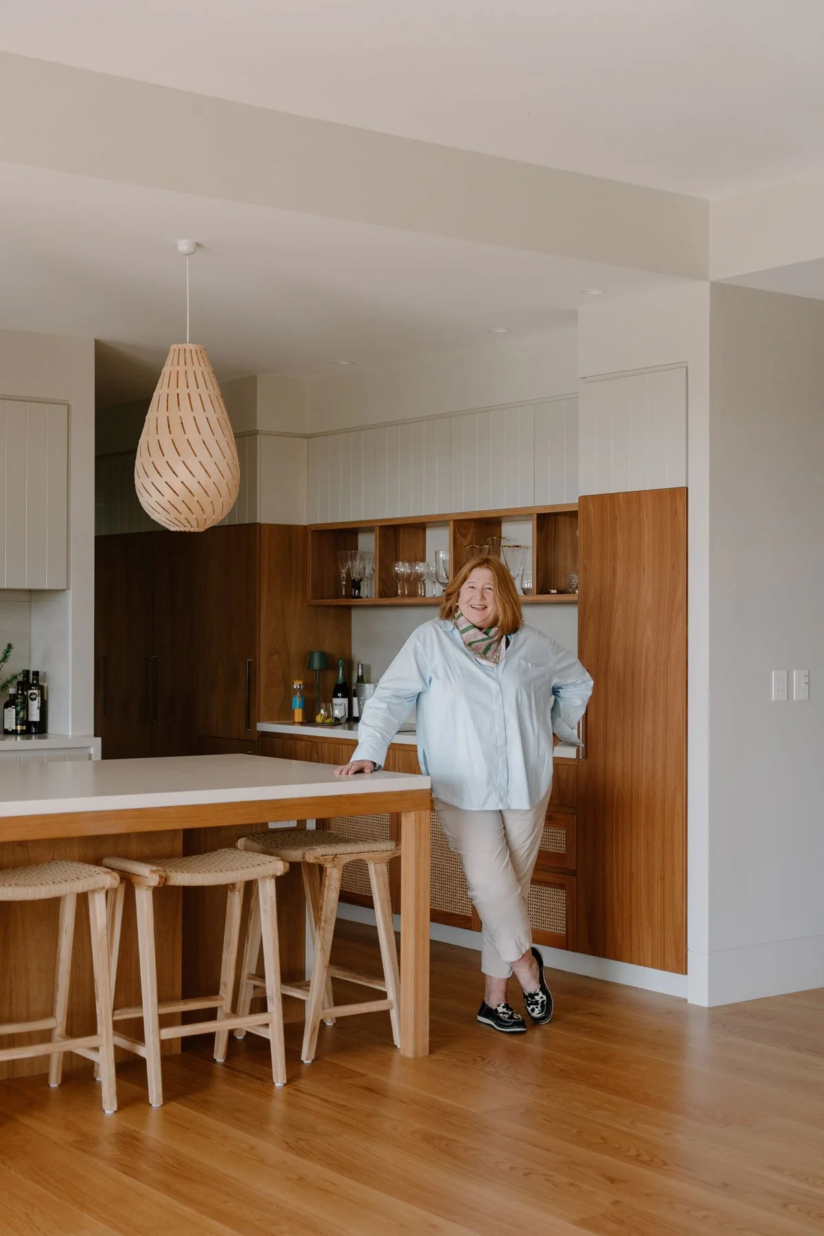 Suzie, smiling and leaning against a kitchen counter in a modern kitchen with wooden cabinets and a white countertop.