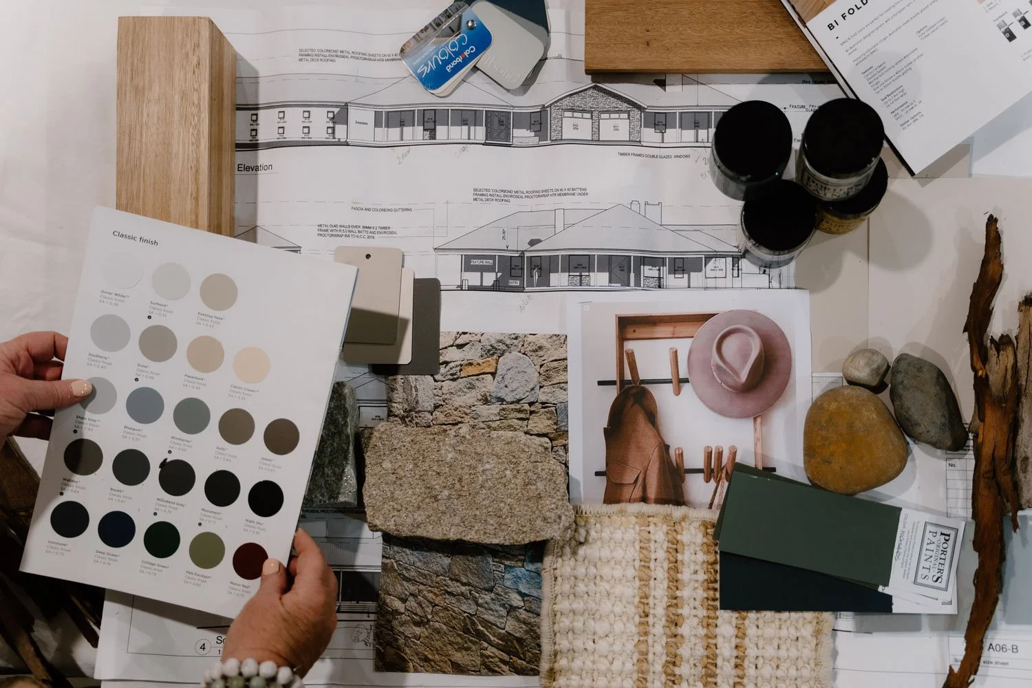 A flat lay of architectural and interior design materials, including paint swatches, stone samples, fabric samples, paint cans, a color palette, design plans, photographs, and wood and stone pieces.
